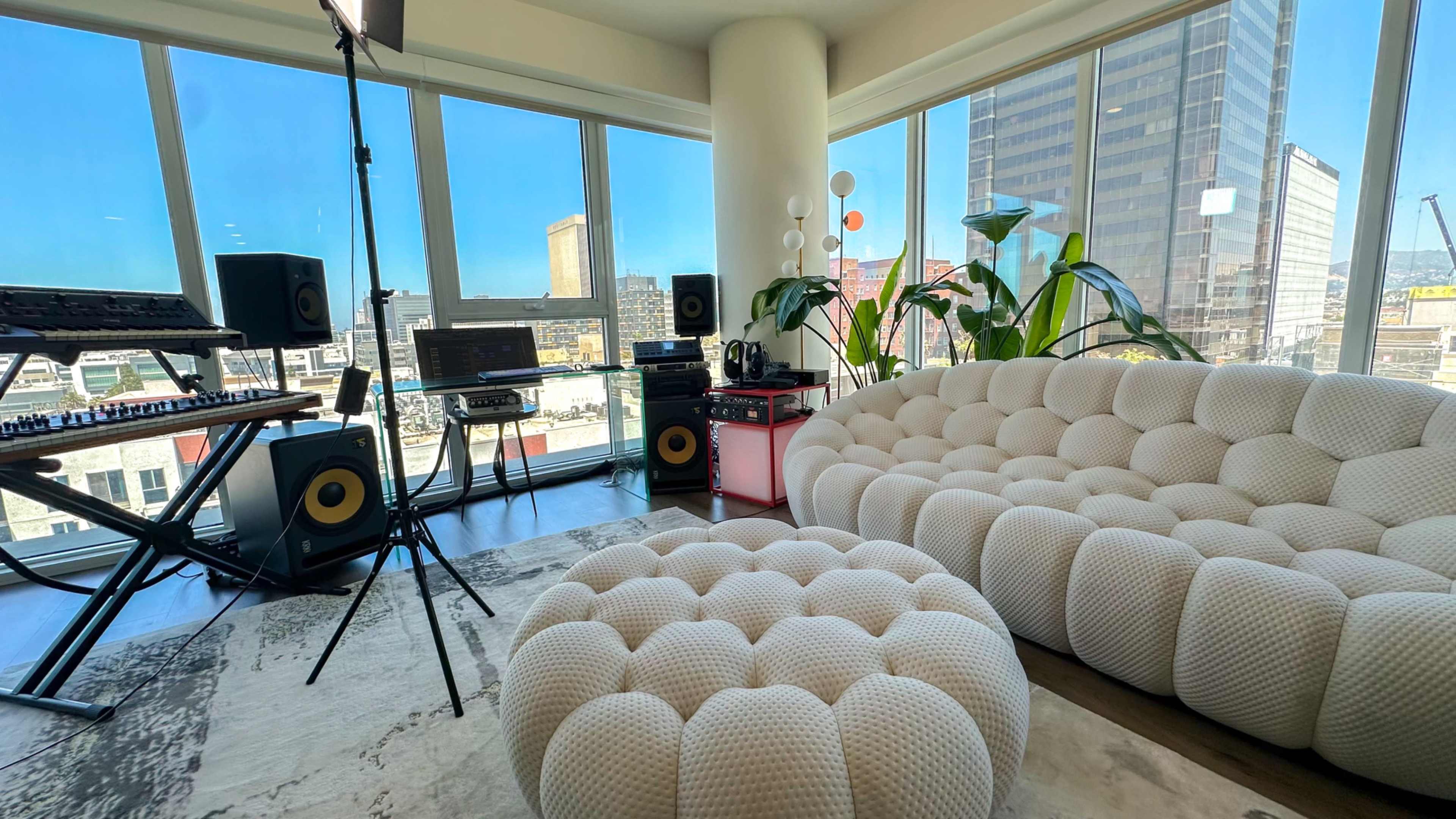 The image shows a modern living room with a large white tufted sofa and ottoman, set against expansive windows providing a view of a city skyline, along with music equipment and plants.
