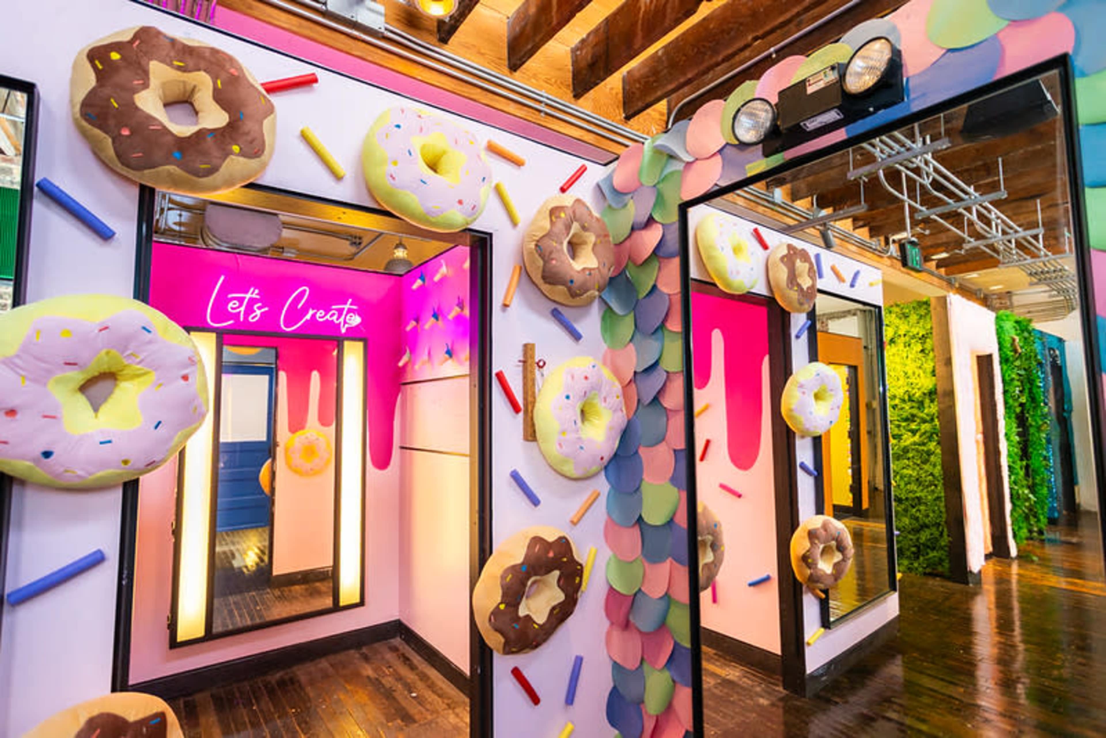 The image shows a colorful room with large donut decorations on the walls and mirrors, accompanied by a vibrant neon sign that reads "Let's Create."