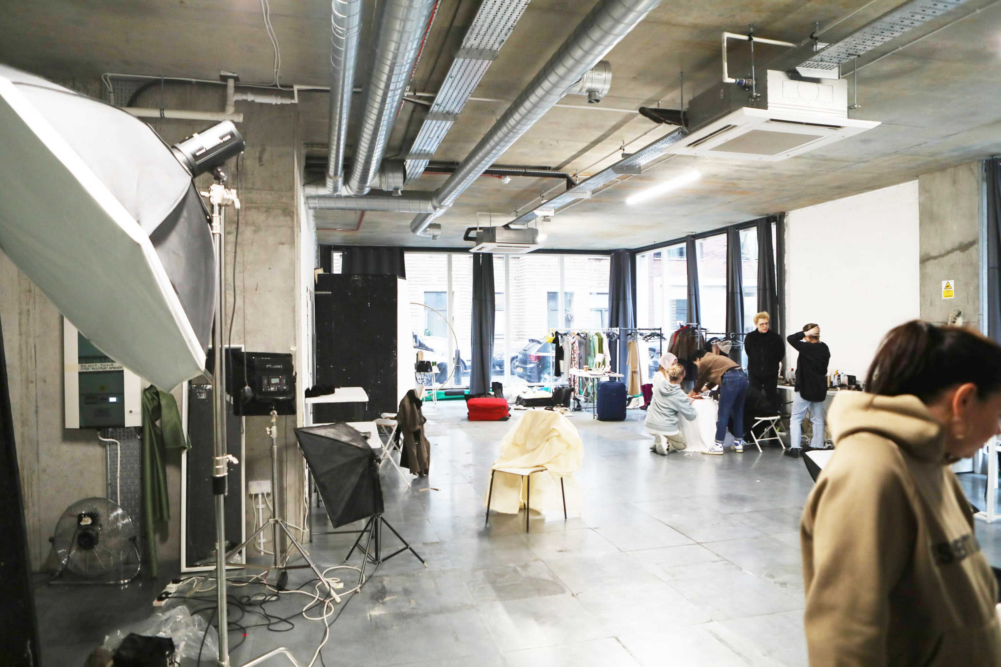 A spacious studio with large windows features clothing racks, a backdrop, and several individuals preparing for a photo shoot.