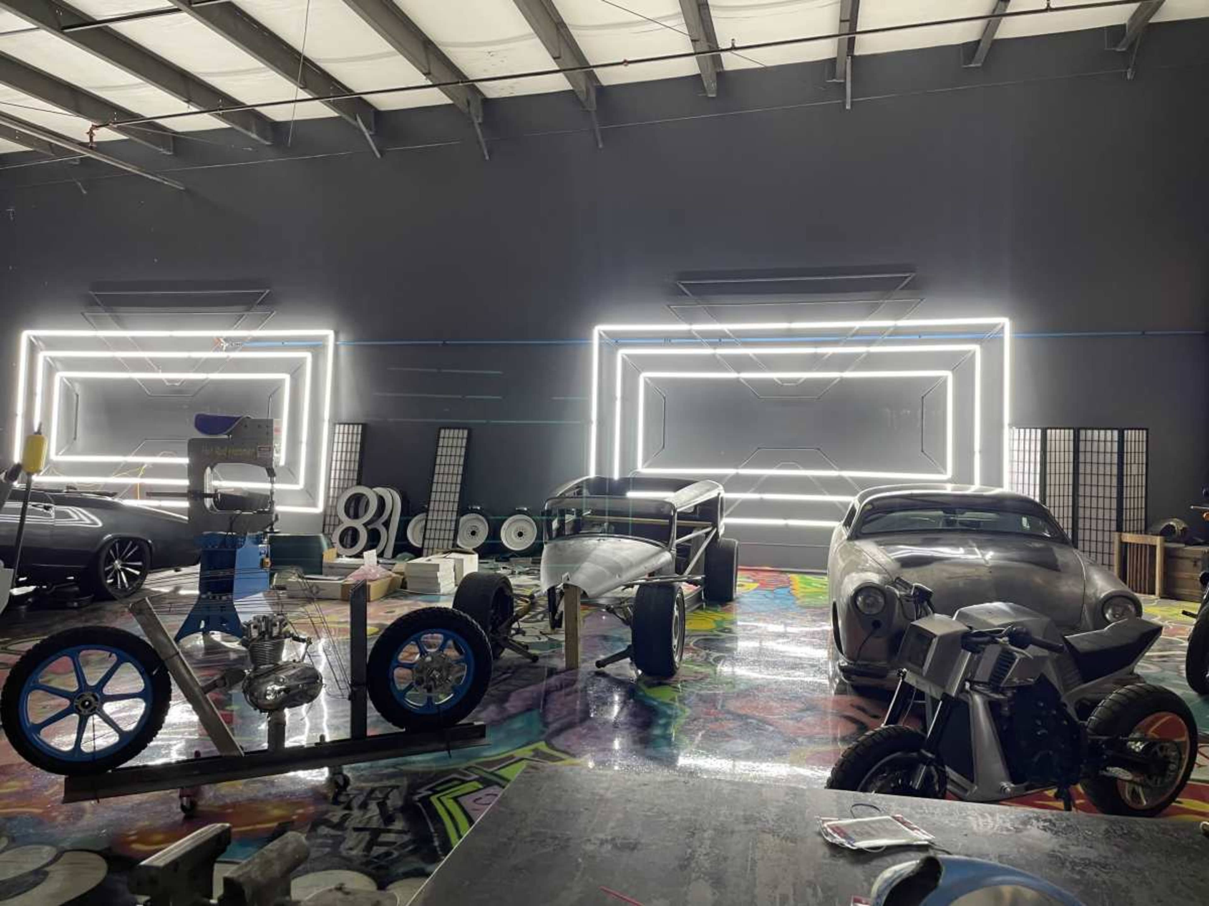 The image shows a warehouse filled with various vehicles and automotive parts, illuminated by neon light frames on the walls.