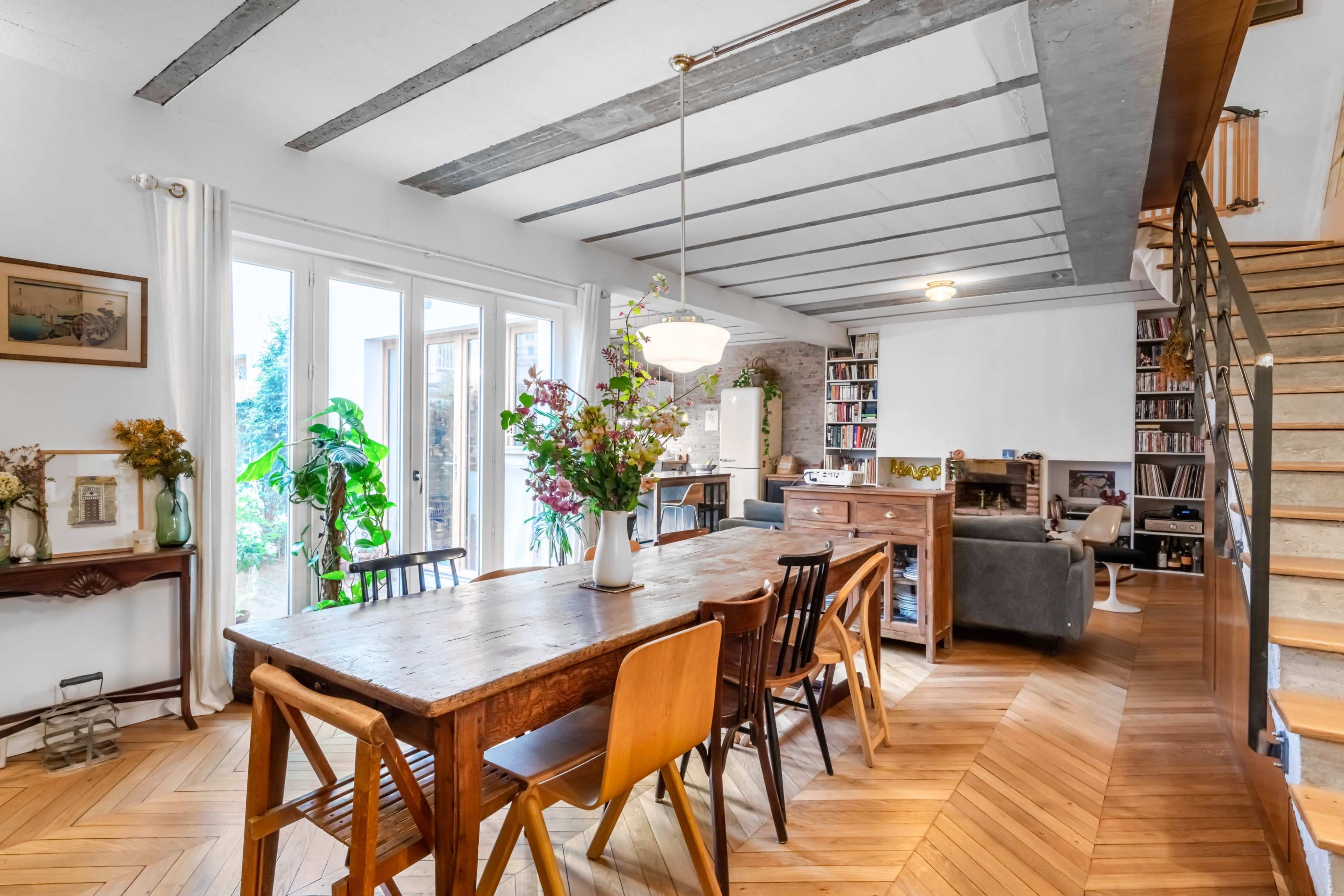 The image shows a bright living space with a wooden dining table surrounded by chairs, large windows, and a staircase leading to a library area.