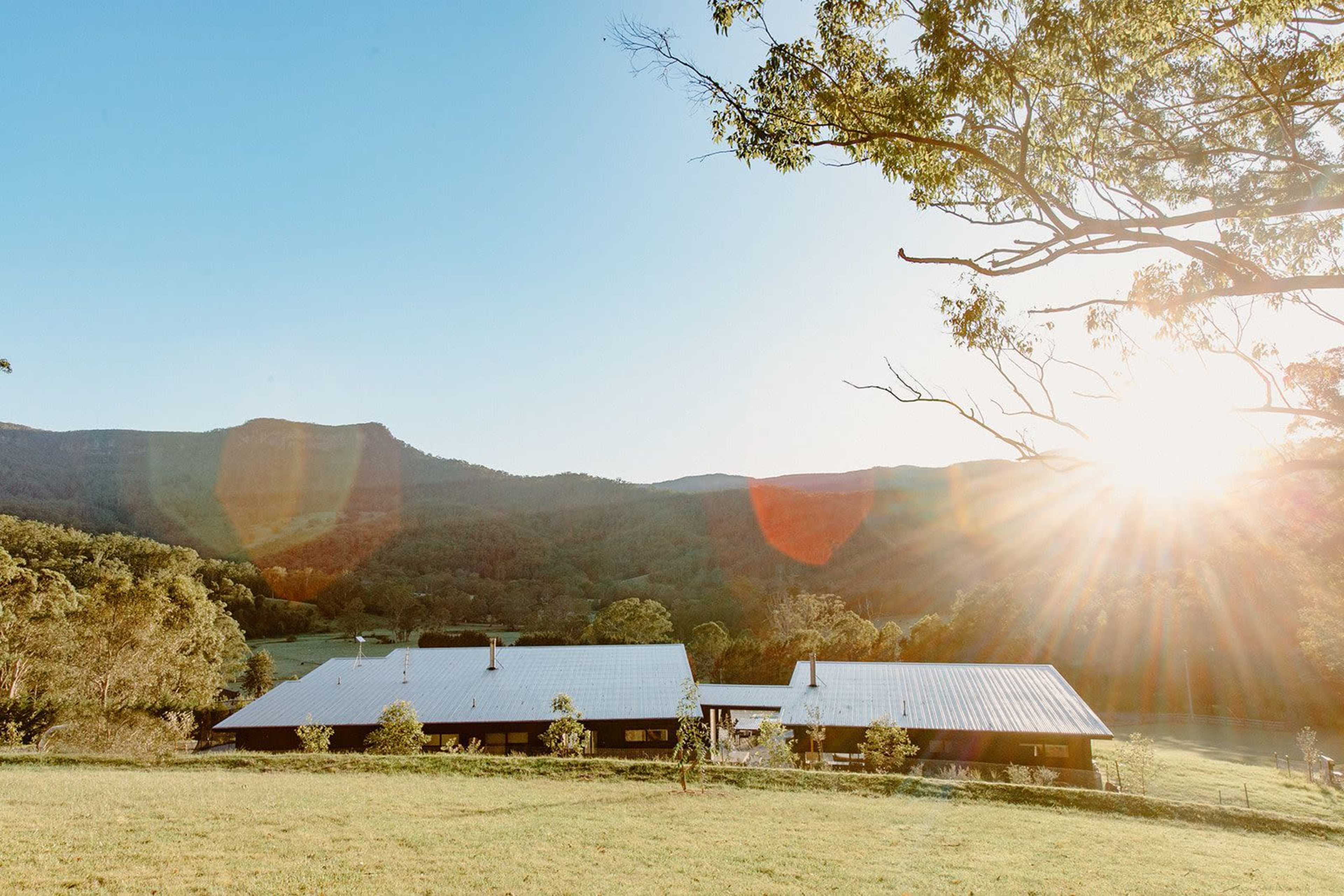 A modern house with a metal roof is situated in a mountainous landscape, illuminated by the setting sun.