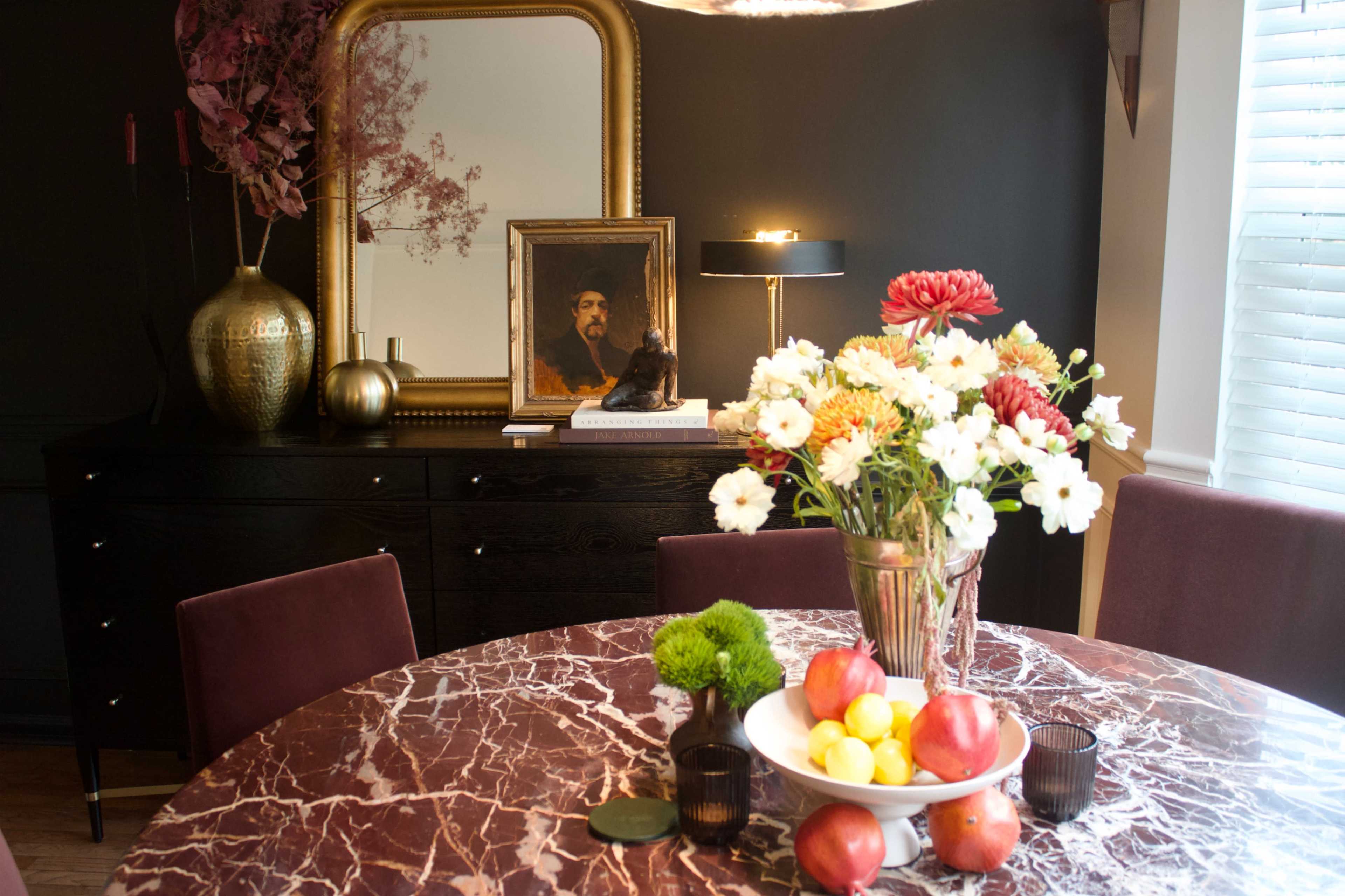 A dining table with a marble surface is adorned with a floral arrangement, fruit, and decorative items, while a framed portrait and a lamp are visible in the background against dark walls.