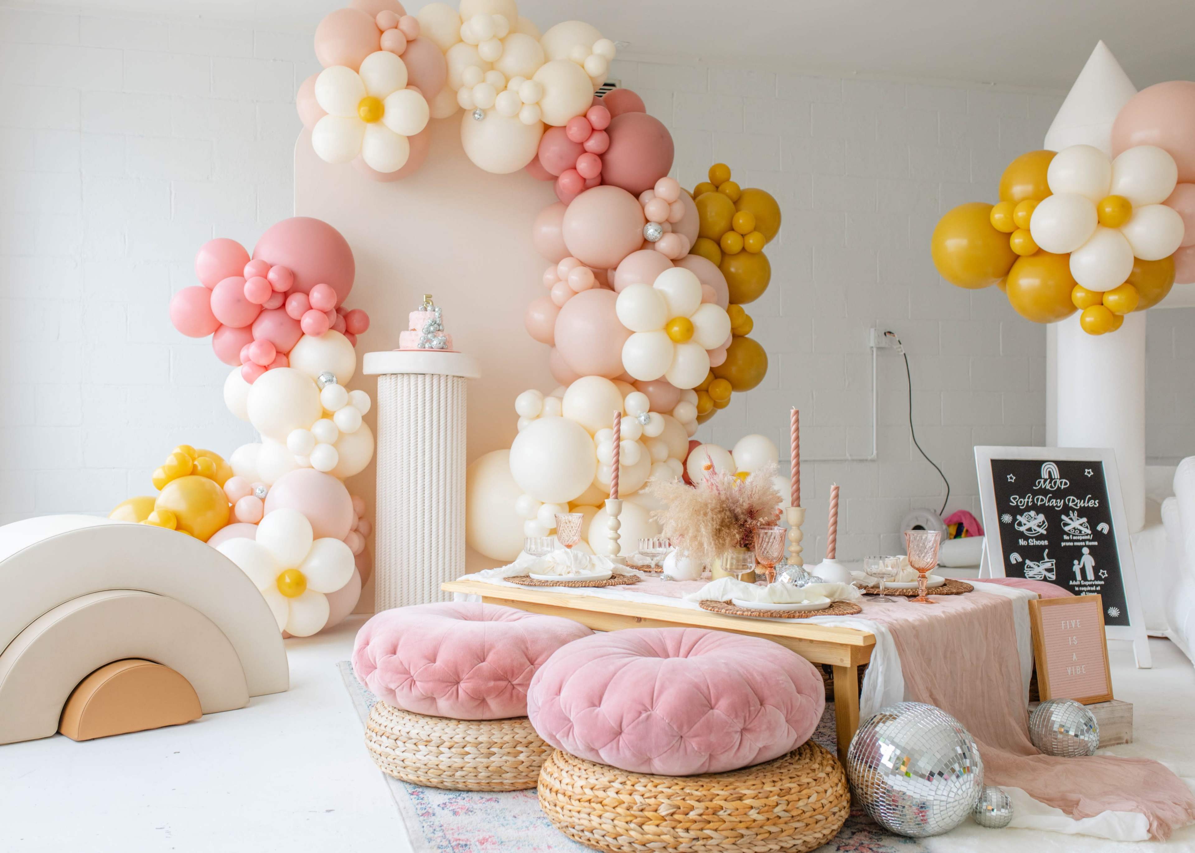 A decorated room featuring a table set with candles and desserts, surrounded by a colorful balloon backdrop and plush seating.