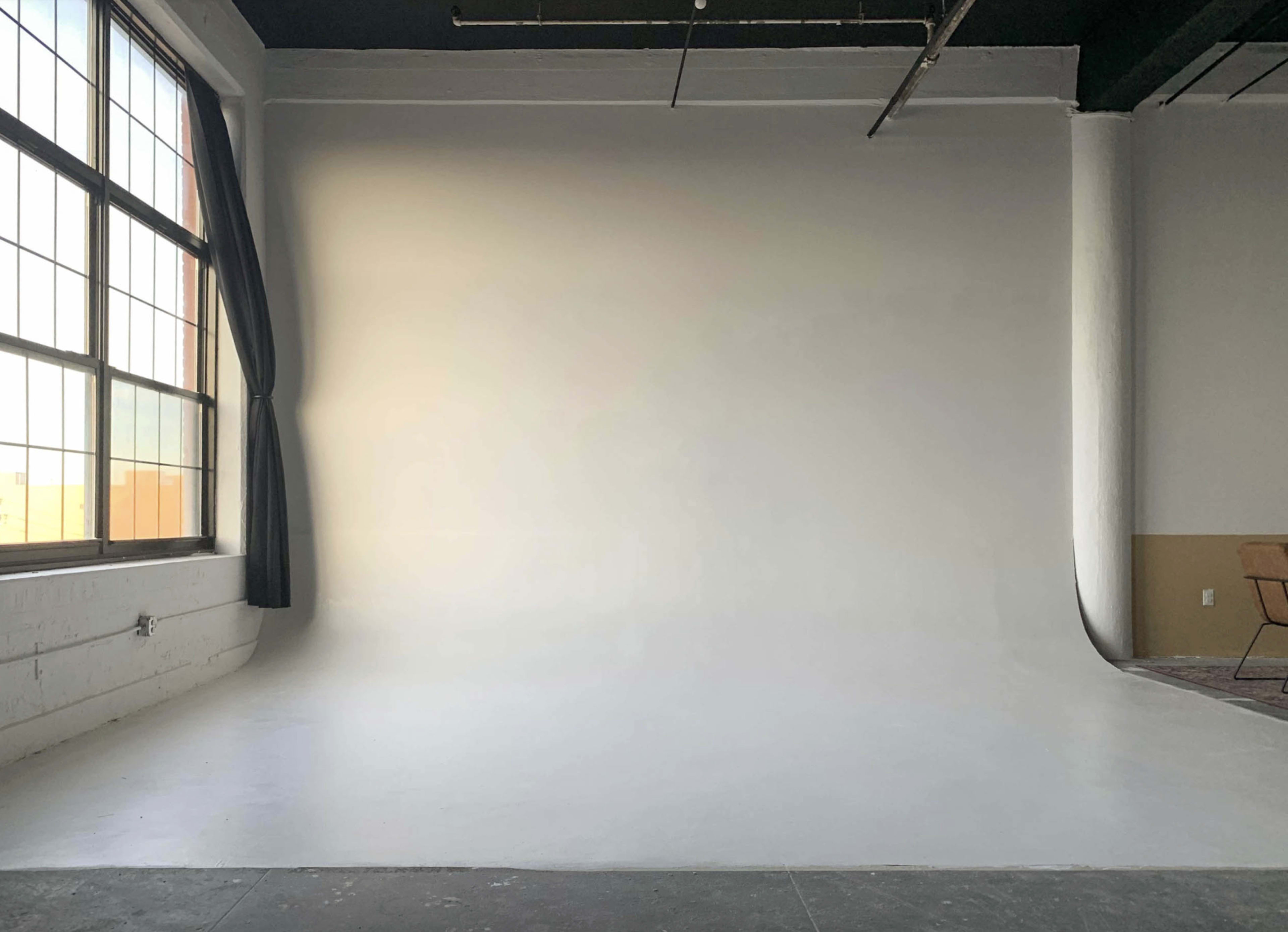 The image shows a minimalist photography studio featuring a large white backdrop, a window with a curtain, and a wooden chair in the corner.