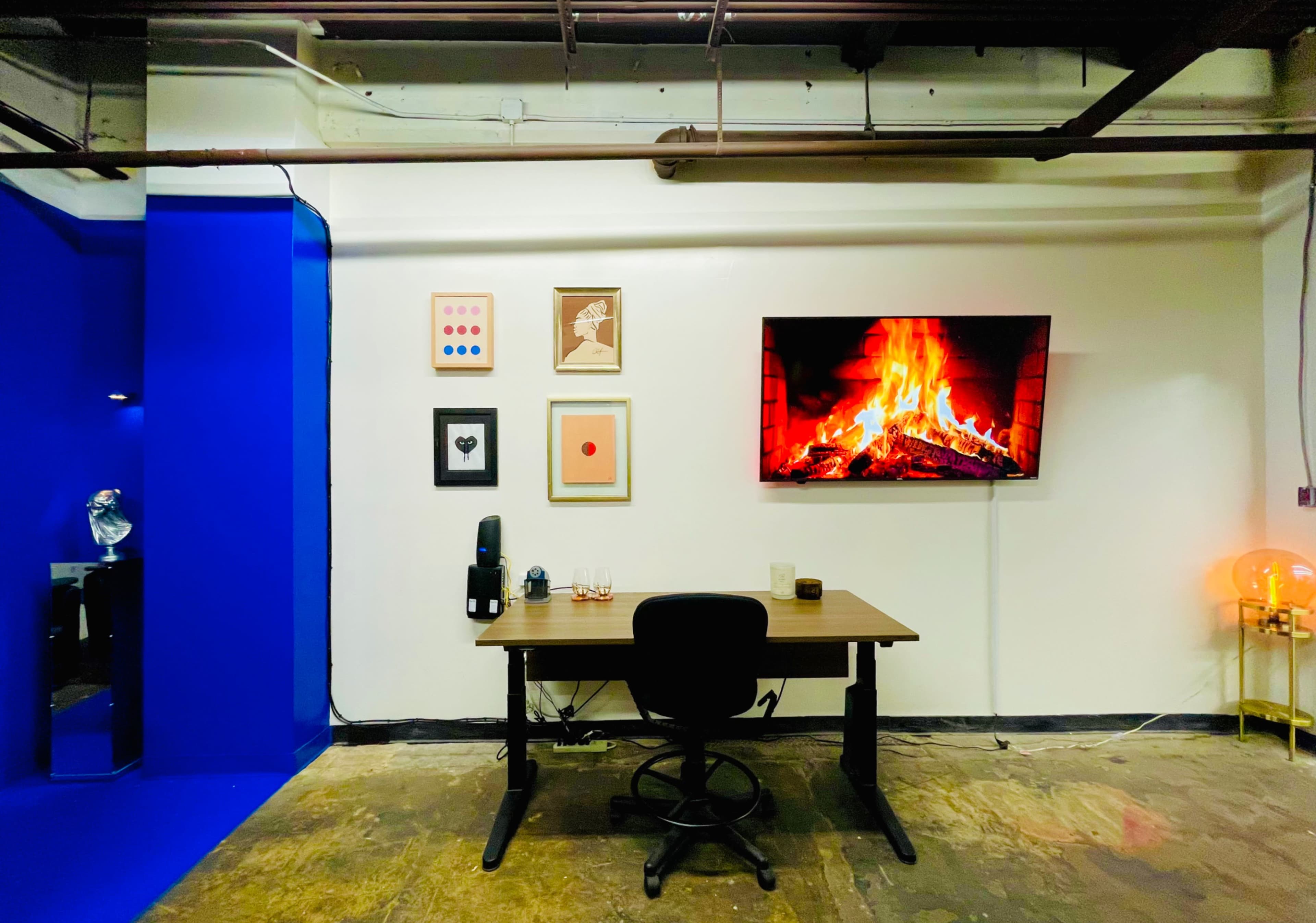 The image shows a minimalistic office space featuring a wooden desk, a television displaying a fireplace, and framed artwork on the wall.