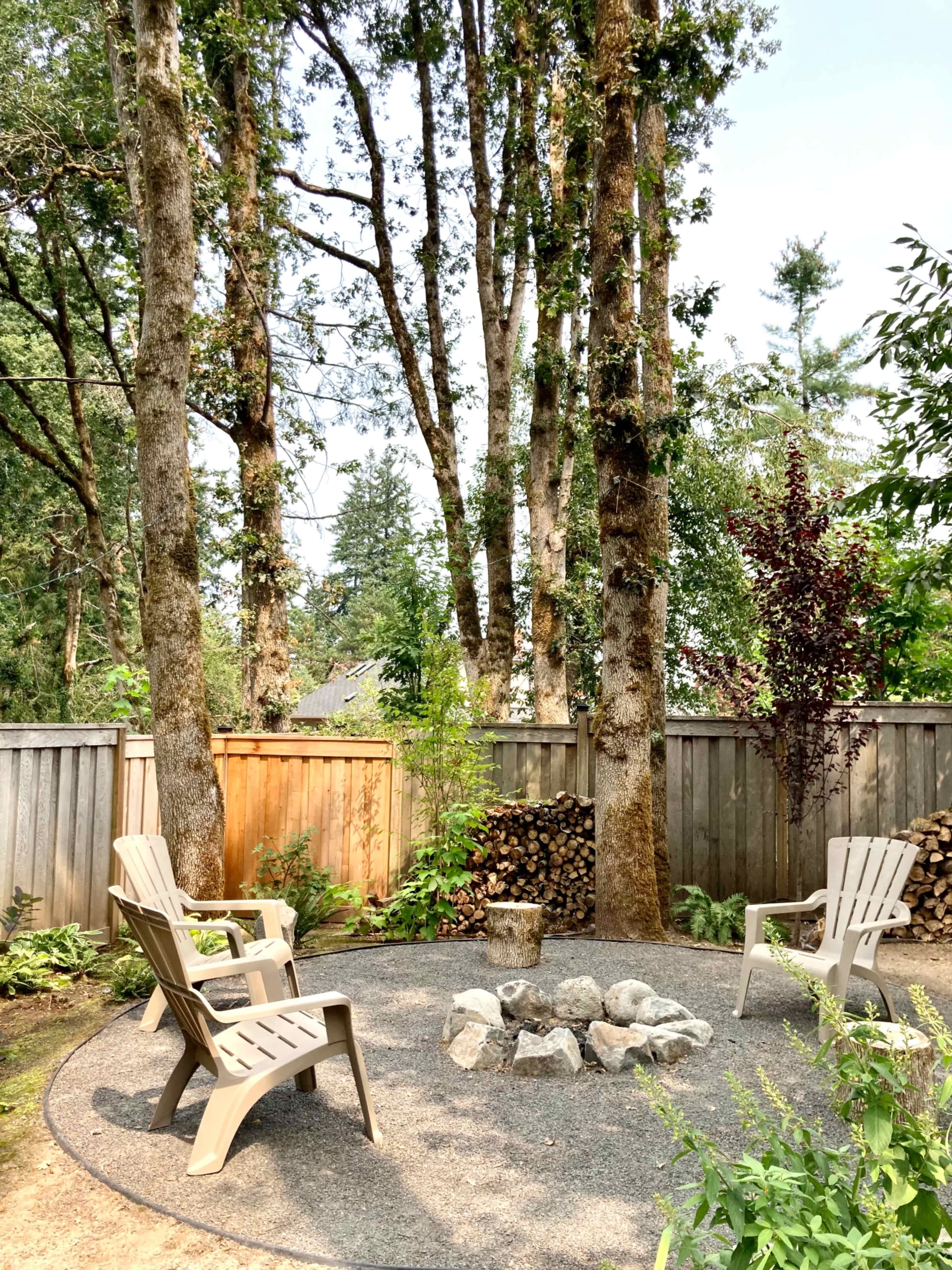 A circular gravel seating area with four chairs around a stone fire pit, surrounded by tall trees and a wooden fence.