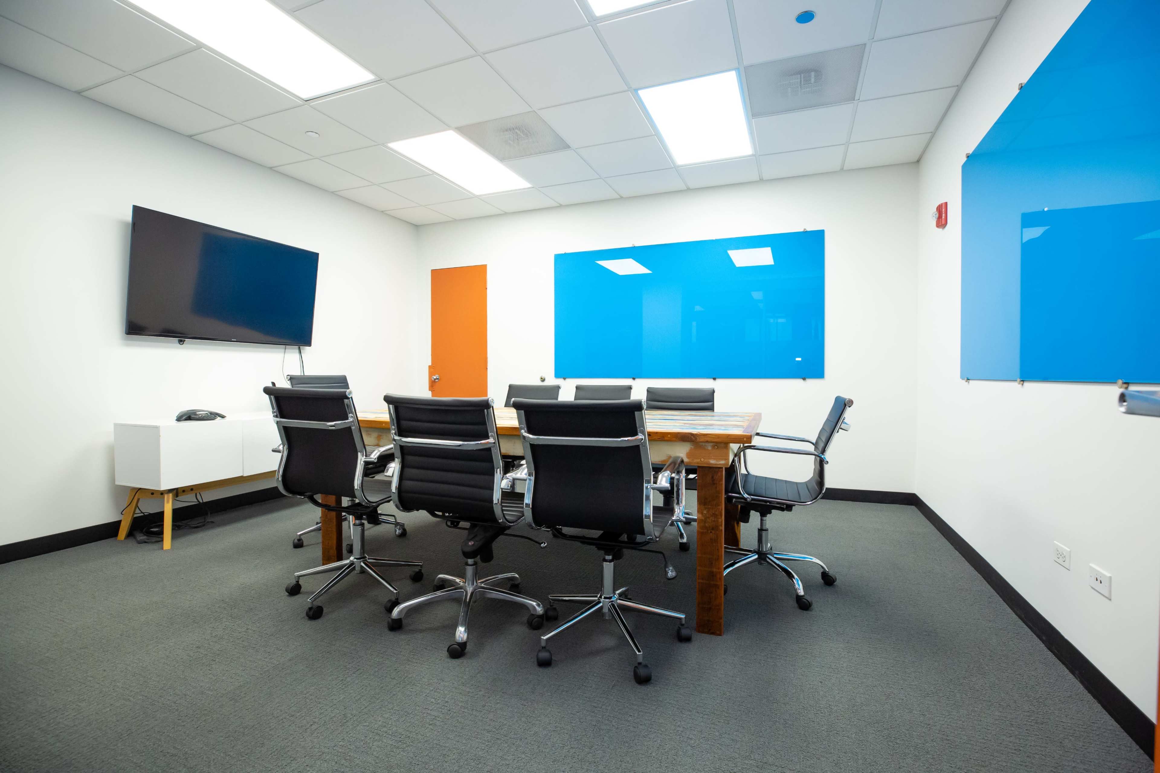 A modern conference room features a long wooden table surrounded by black ergonomic chairs, a large television on the wall, and blue glass boards.