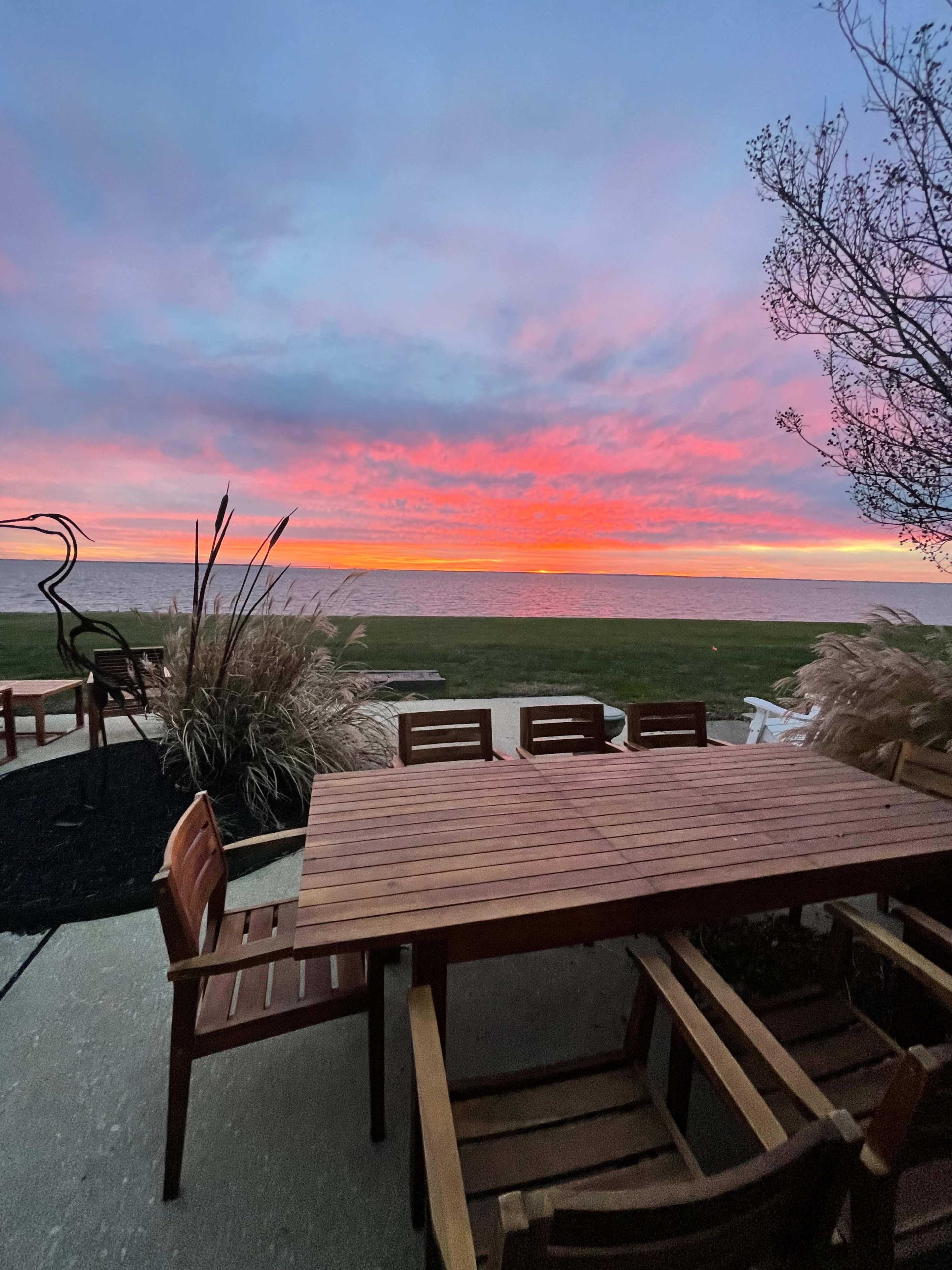A wooden dining table and chairs are set on a patio overlooking a vibrant sunset over the water.