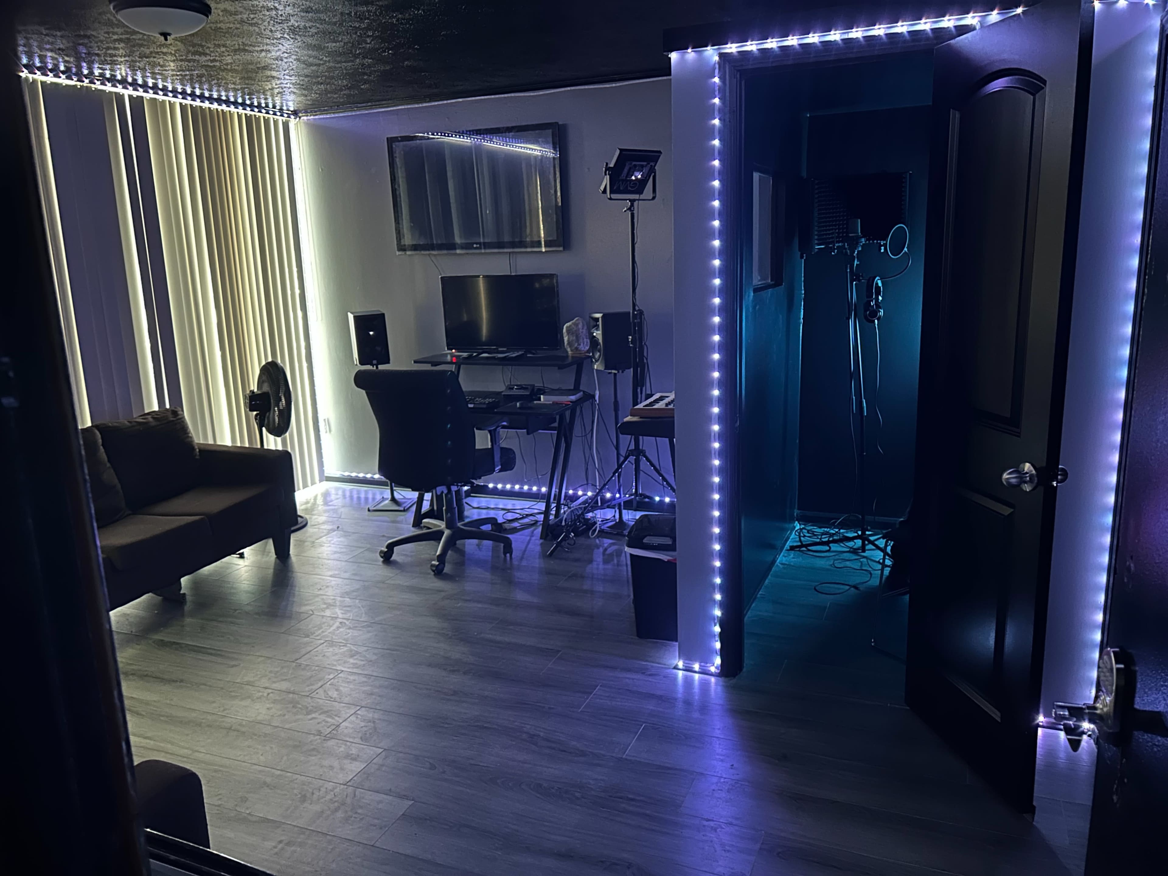A small room with a workspace featuring a desk and computer, enclosed by LED lights, leading to a recording area with sound equipment.
