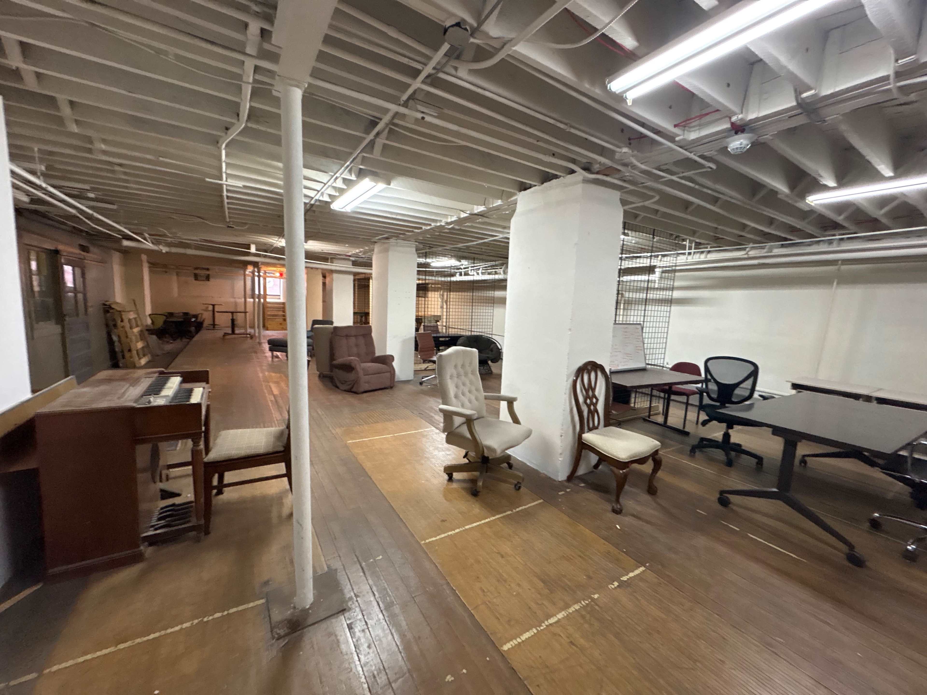 The image shows a large, empty basement room with wooden flooring, several chairs, a desk, and a piano against the wall.