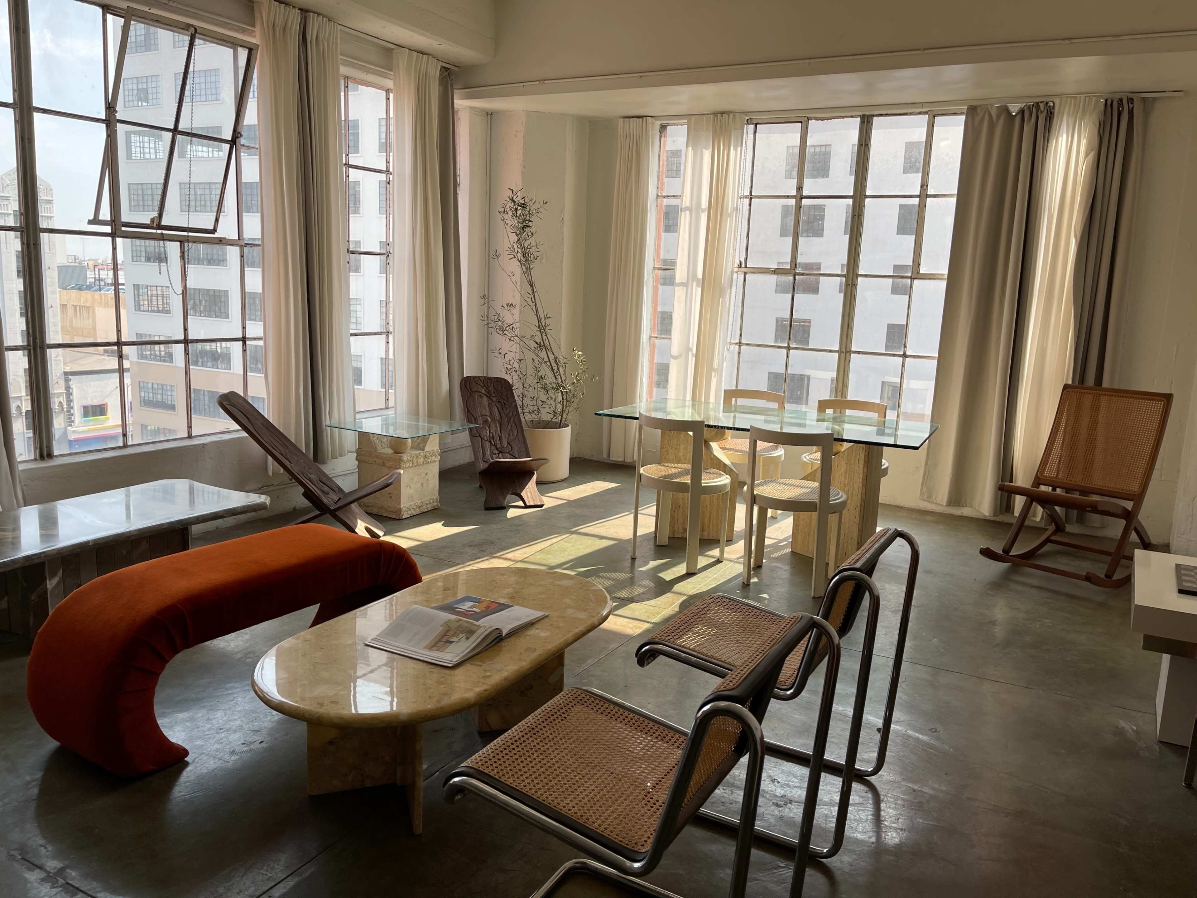 A sunlit room with large windows, featuring modern furniture including a curved red bench, a marble table, and a glass dining table with wooden chairs.