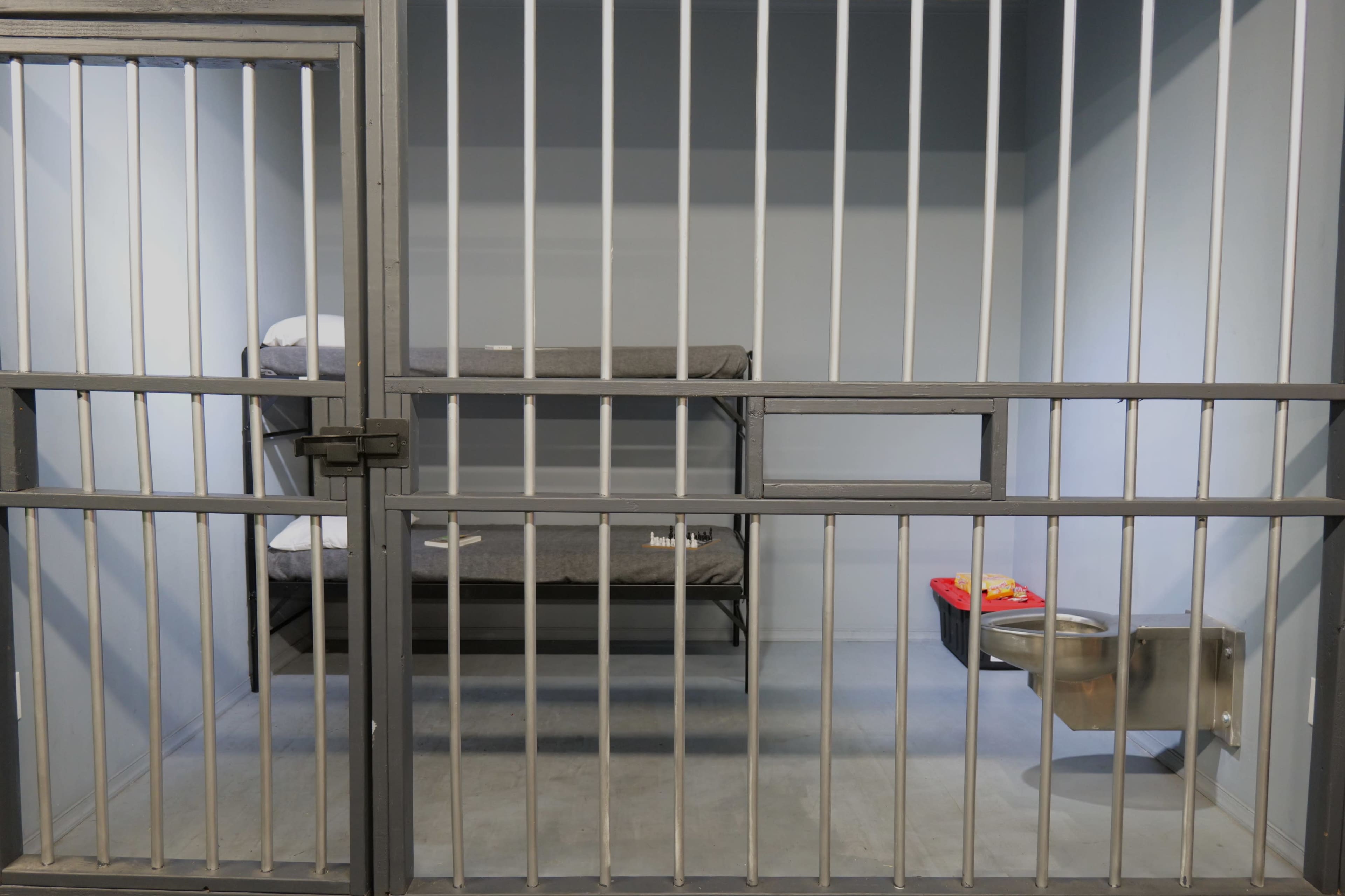 A bare jail cell with metal bars, two beds, and a toilet.