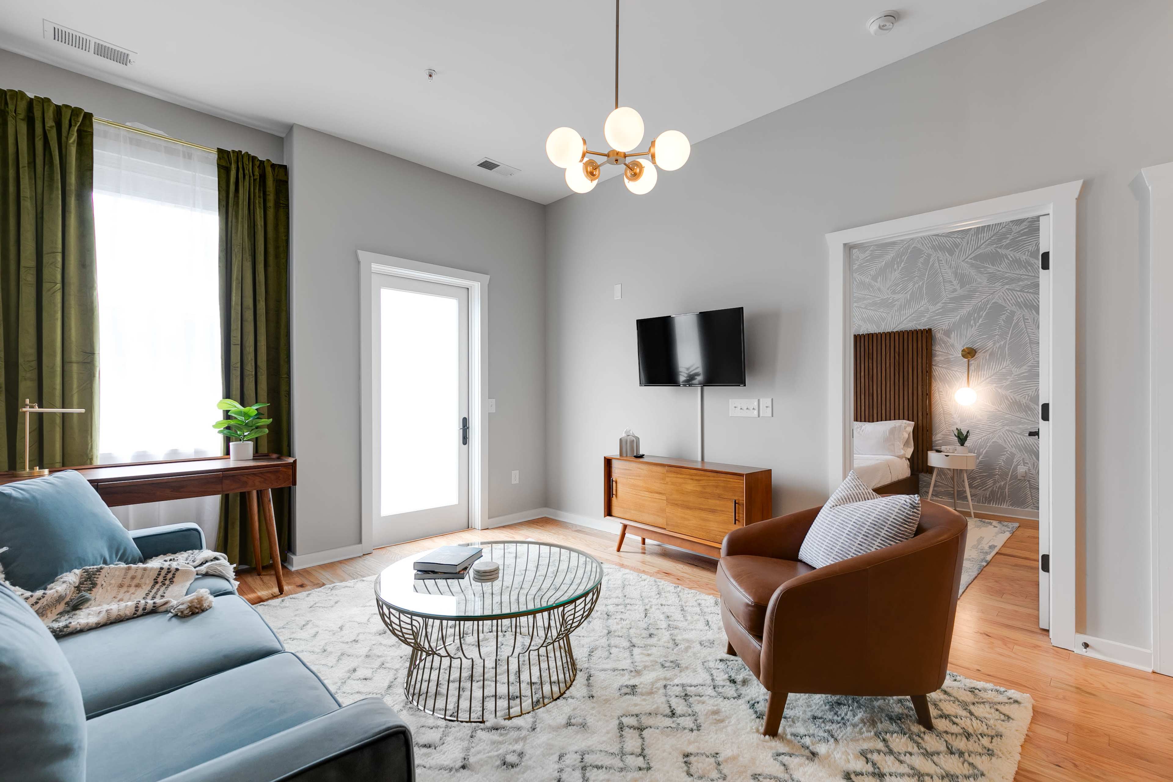 The living room features a blue sofa, a brown leather armchair, a round coffee table, and a television mounted on the wall, with green curtains and a plant by the window.