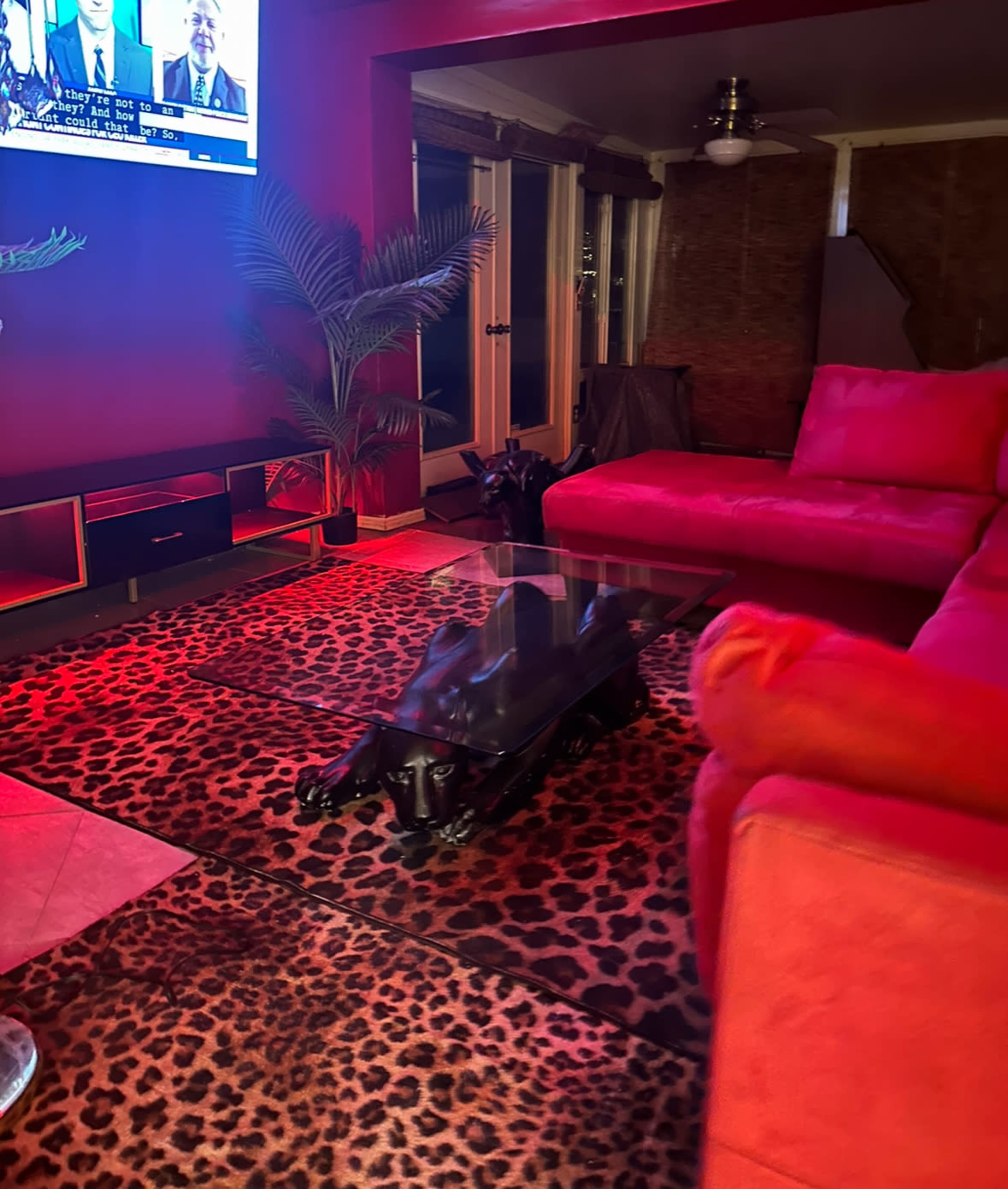 A living room features a red sectional couch, a glass coffee table on a leopard print rug, and a large TV displaying a news broadcast.