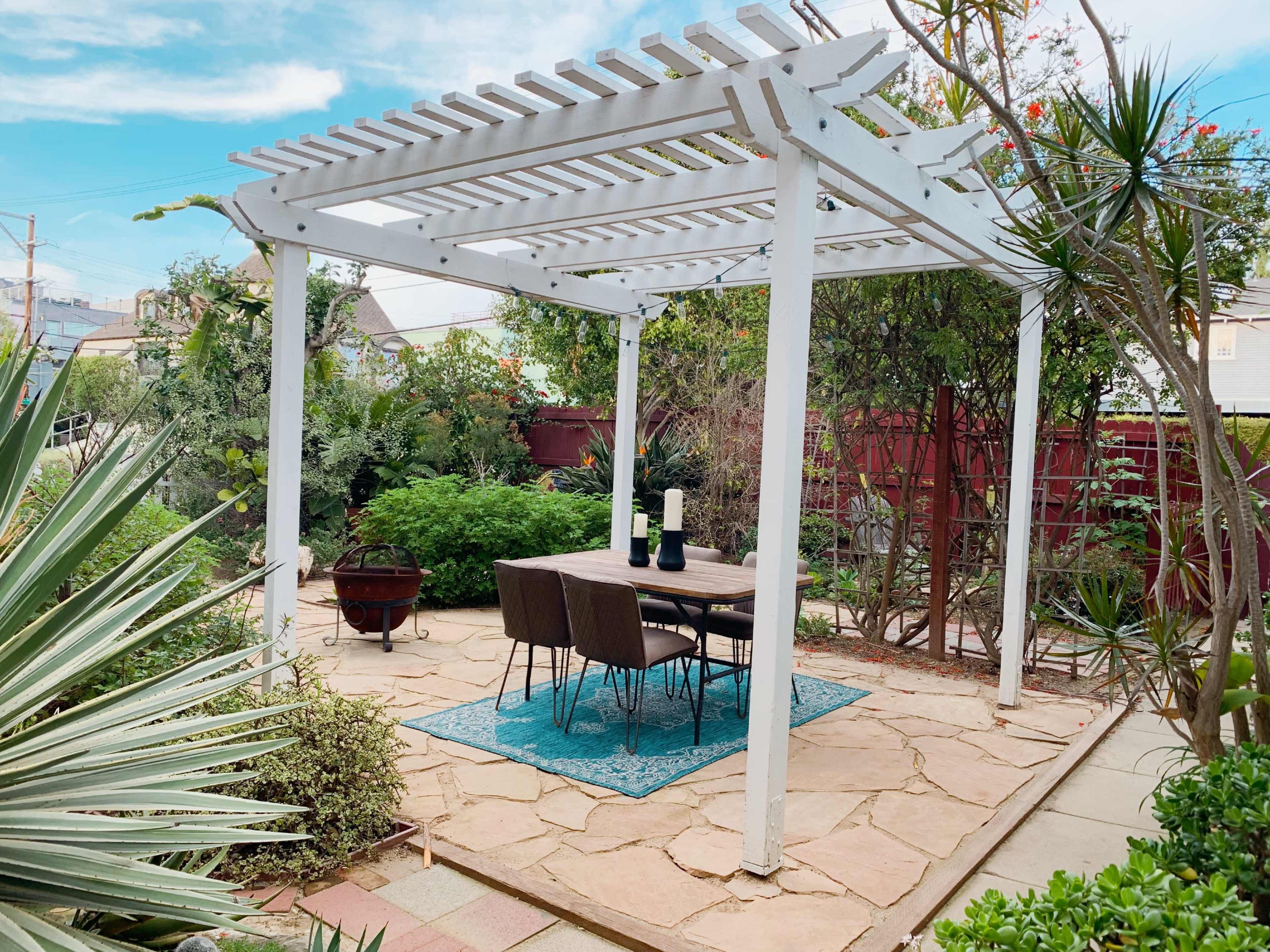 The image shows a patio area with a white pergola, a wooden table set with chairs, and surrounded by lush greenery and stone pathways.