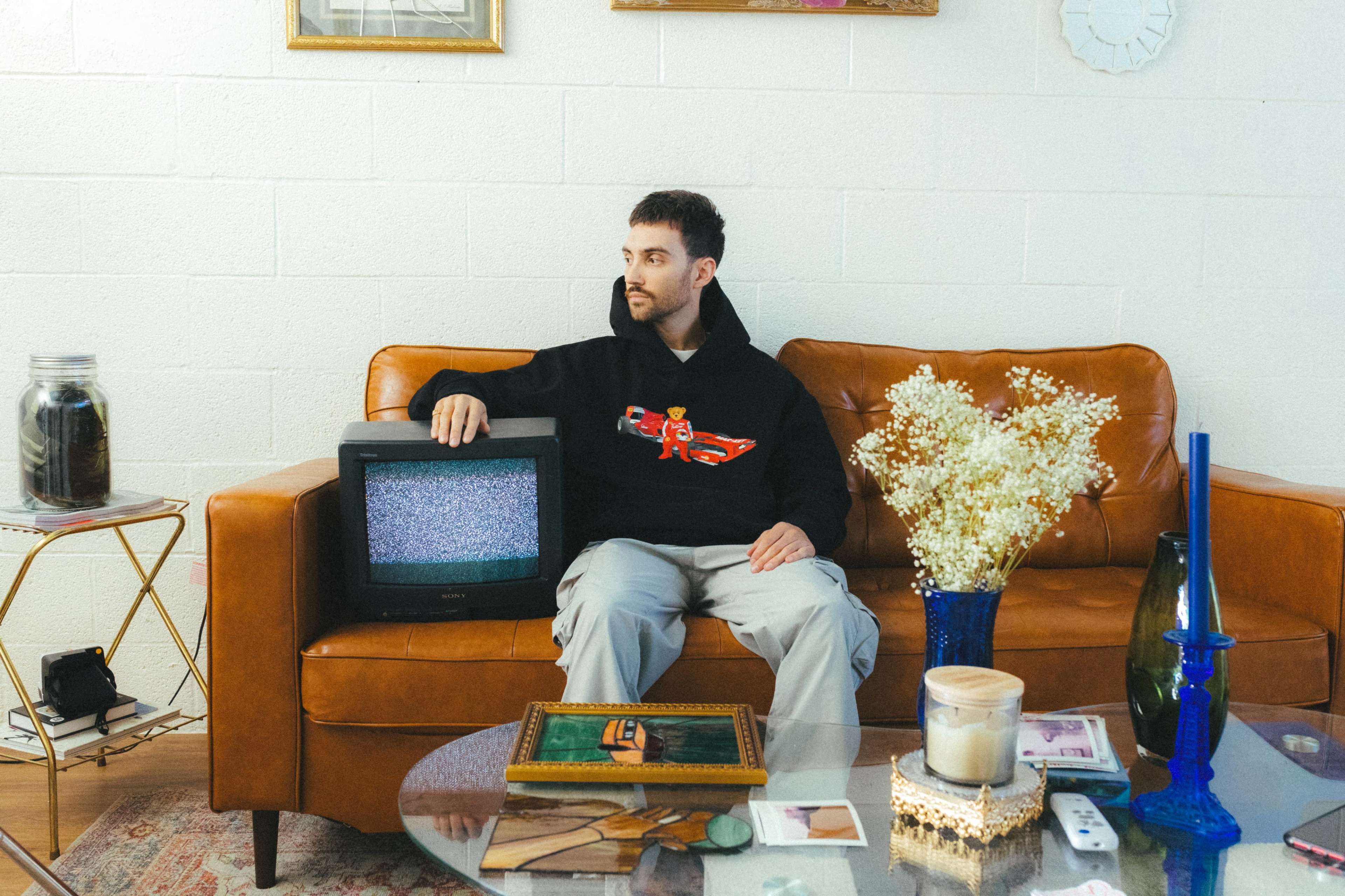 A man in a black hoodie sits on a brown leather couch next to an old television set, surrounded by various decorative items on a coffee table.