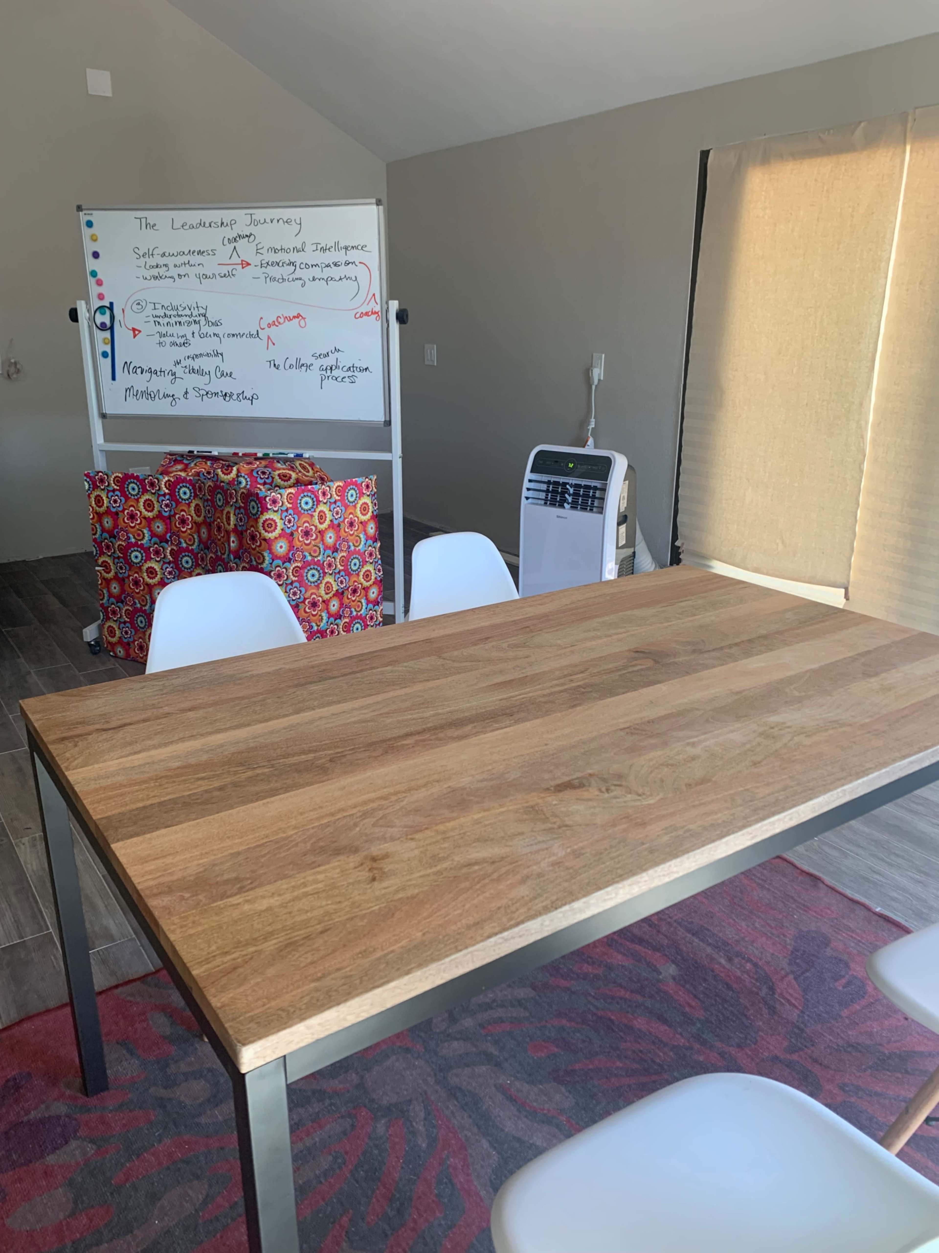 A wooden table with metal legs is positioned in a room featuring a whiteboard covered in notes, a colorful storage cart, and white chairs.