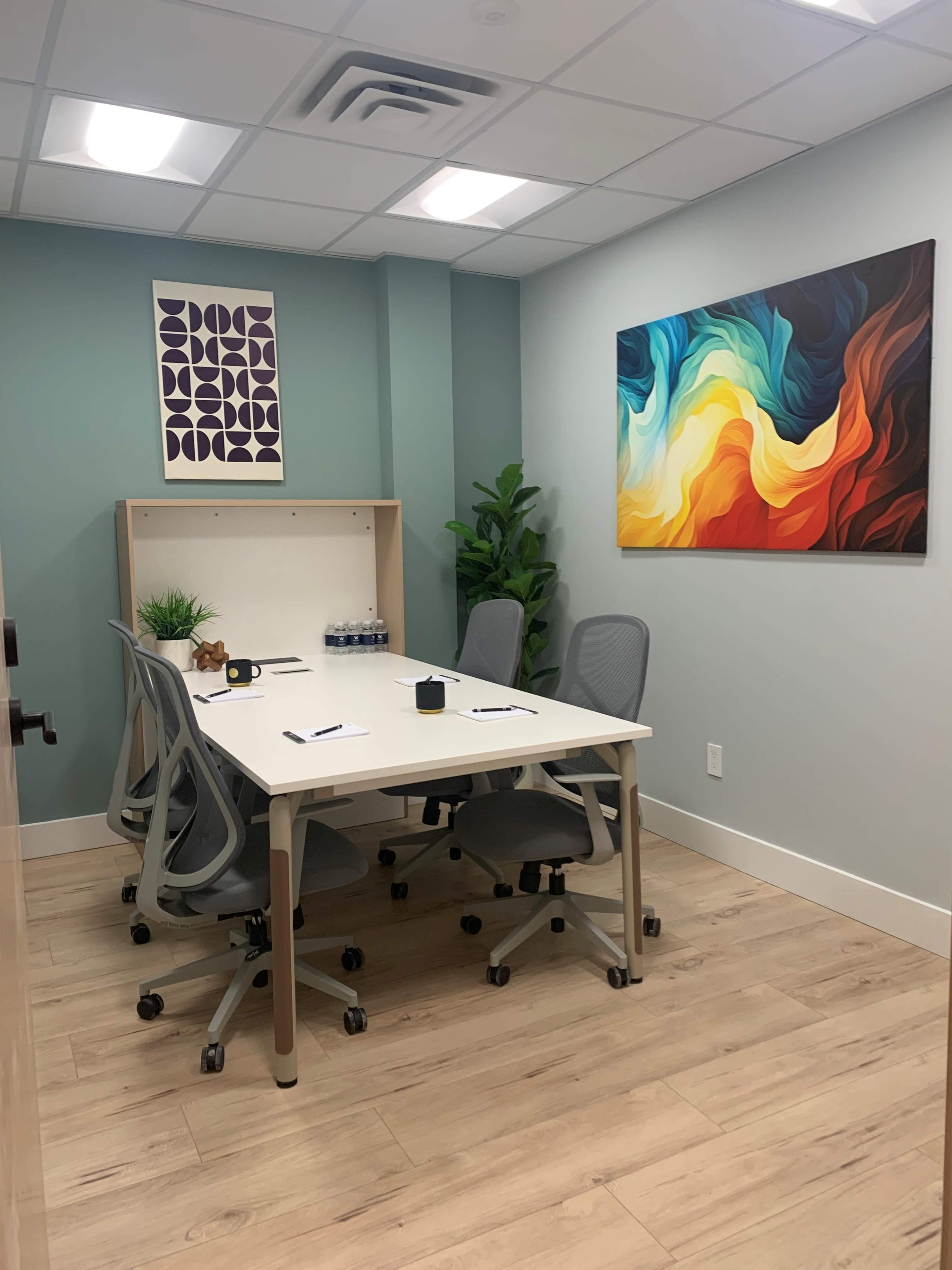 A small conference room features a white table surrounded by four gray chairs, a large colorful abstract painting on one wall, and a plant in the corner.