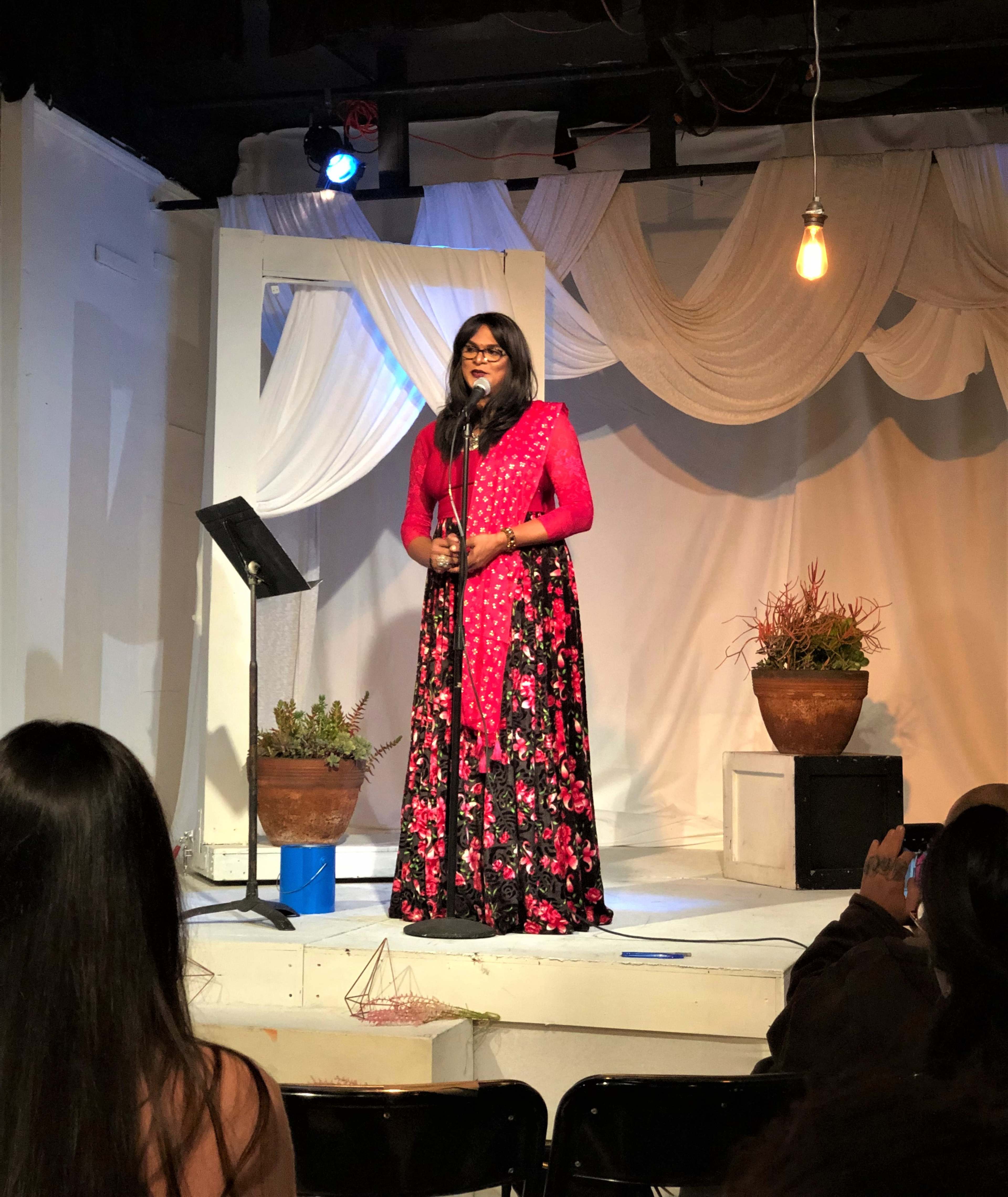 A performer stands on stage in a floral dress, holding a microphone while facing an audience seated in front of her.