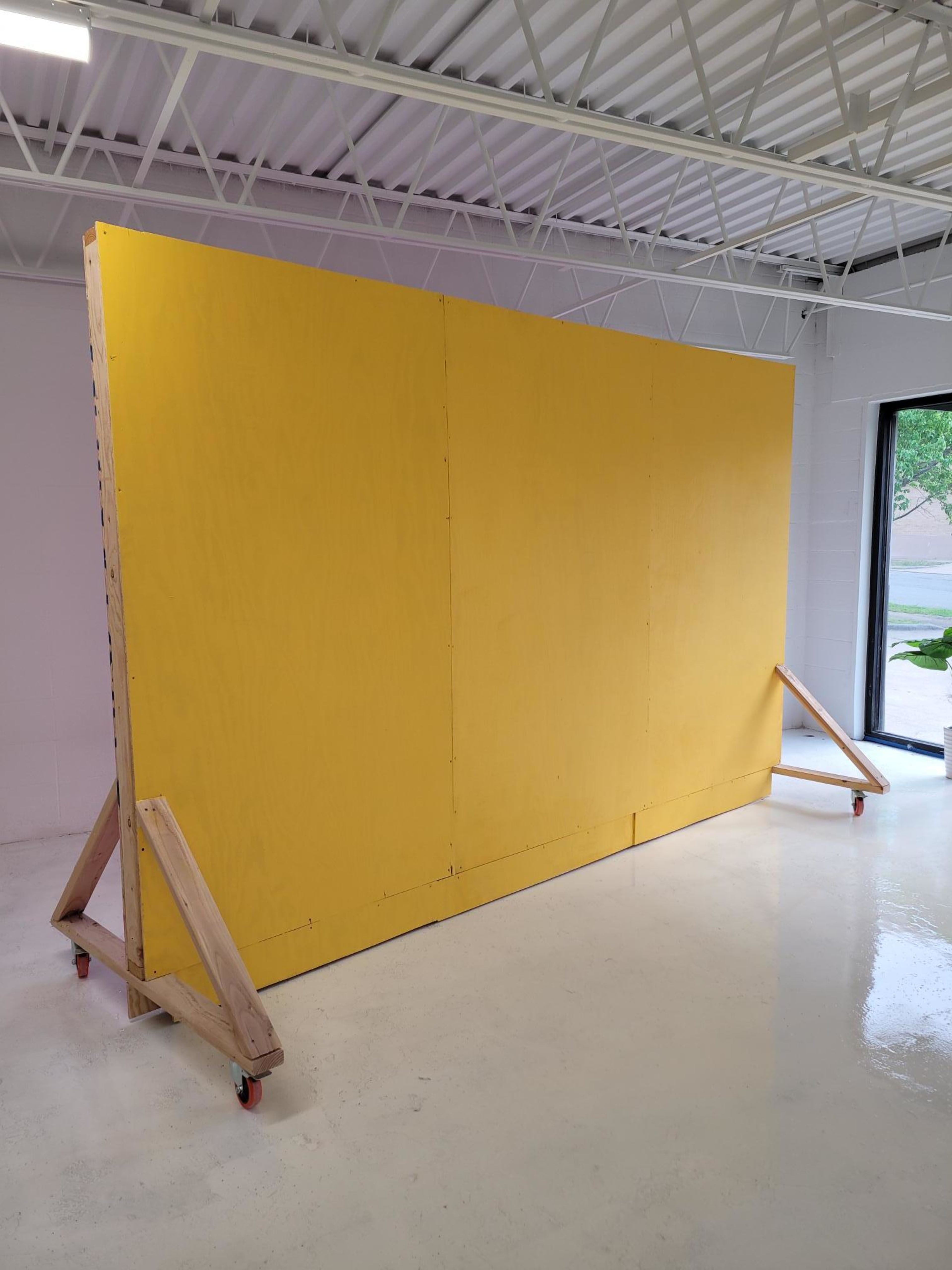 A large yellow wall on wheels is positioned in a bright, empty room with a window.