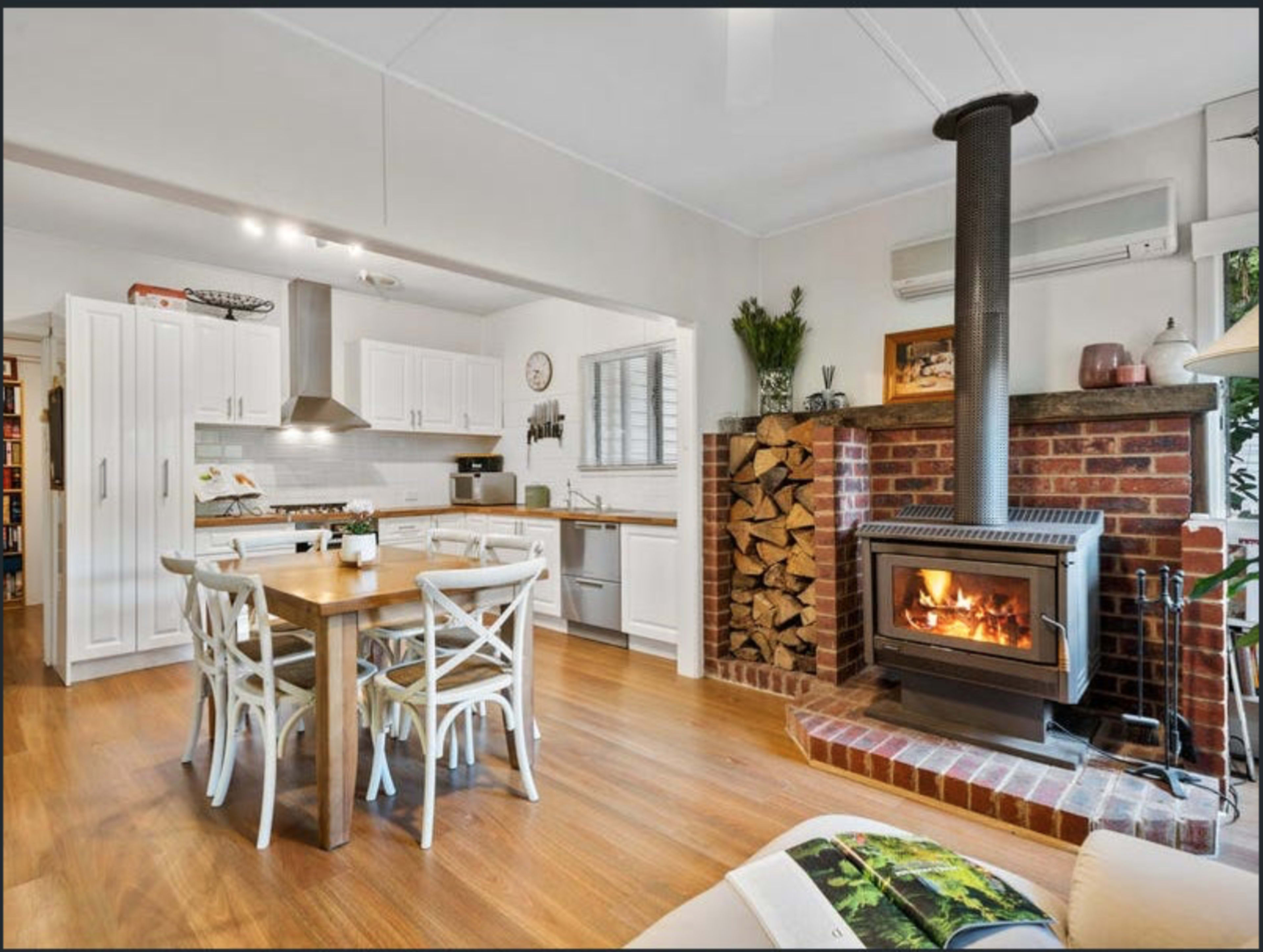 A cozy kitchen and dining area featuring a wooden table with chairs, a brick fireplace with a wood stack beside it, and modern appliances in a bright, open layout.