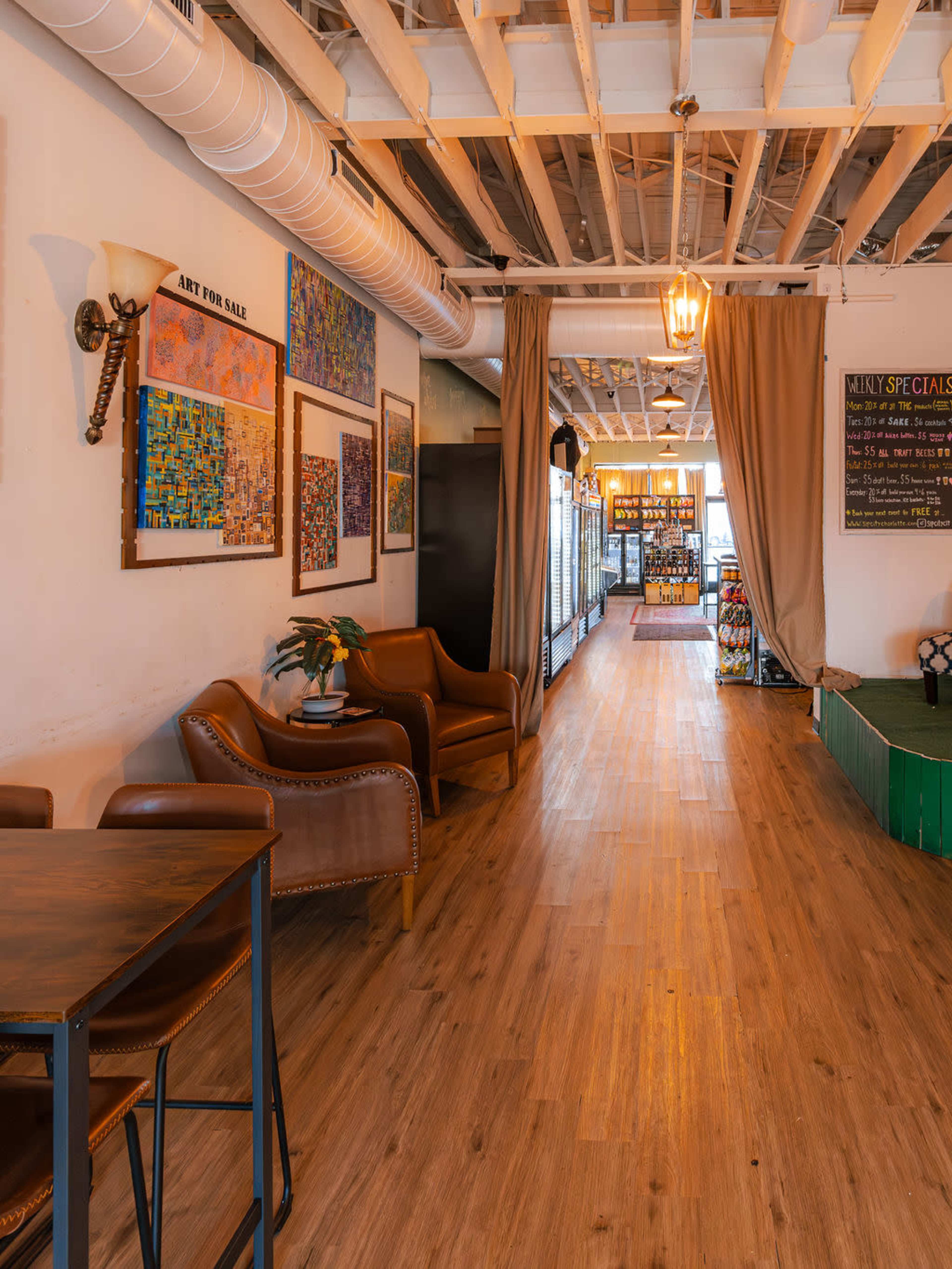 The image shows a cozy interior of a shop or café with wooden flooring, leather chairs, and artwork displayed on the walls, leading to a lighted area with shelves and a chalkboard menu.