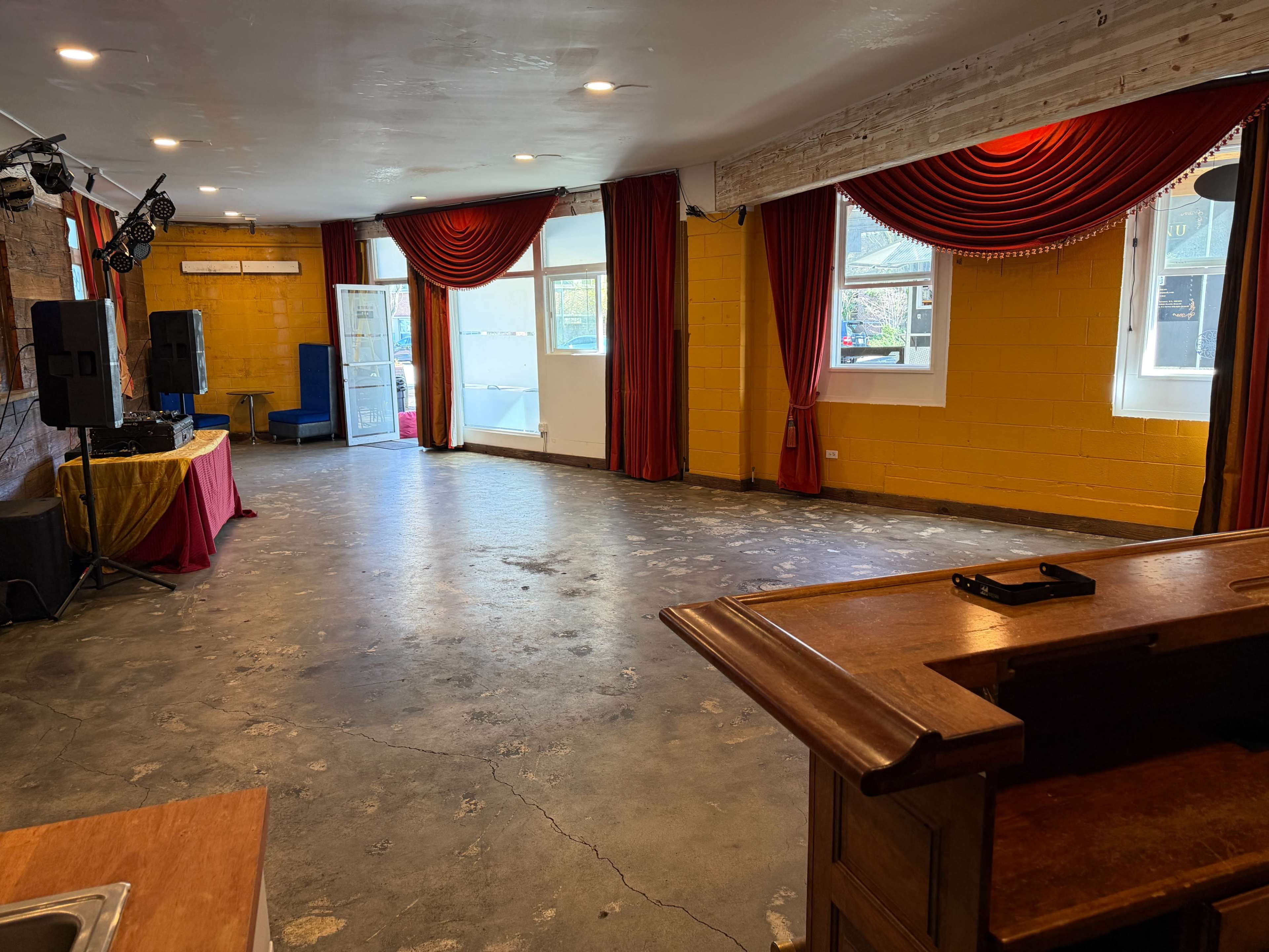The image shows an empty performance space with concrete flooring, red drapes, and a small stage area on one side.