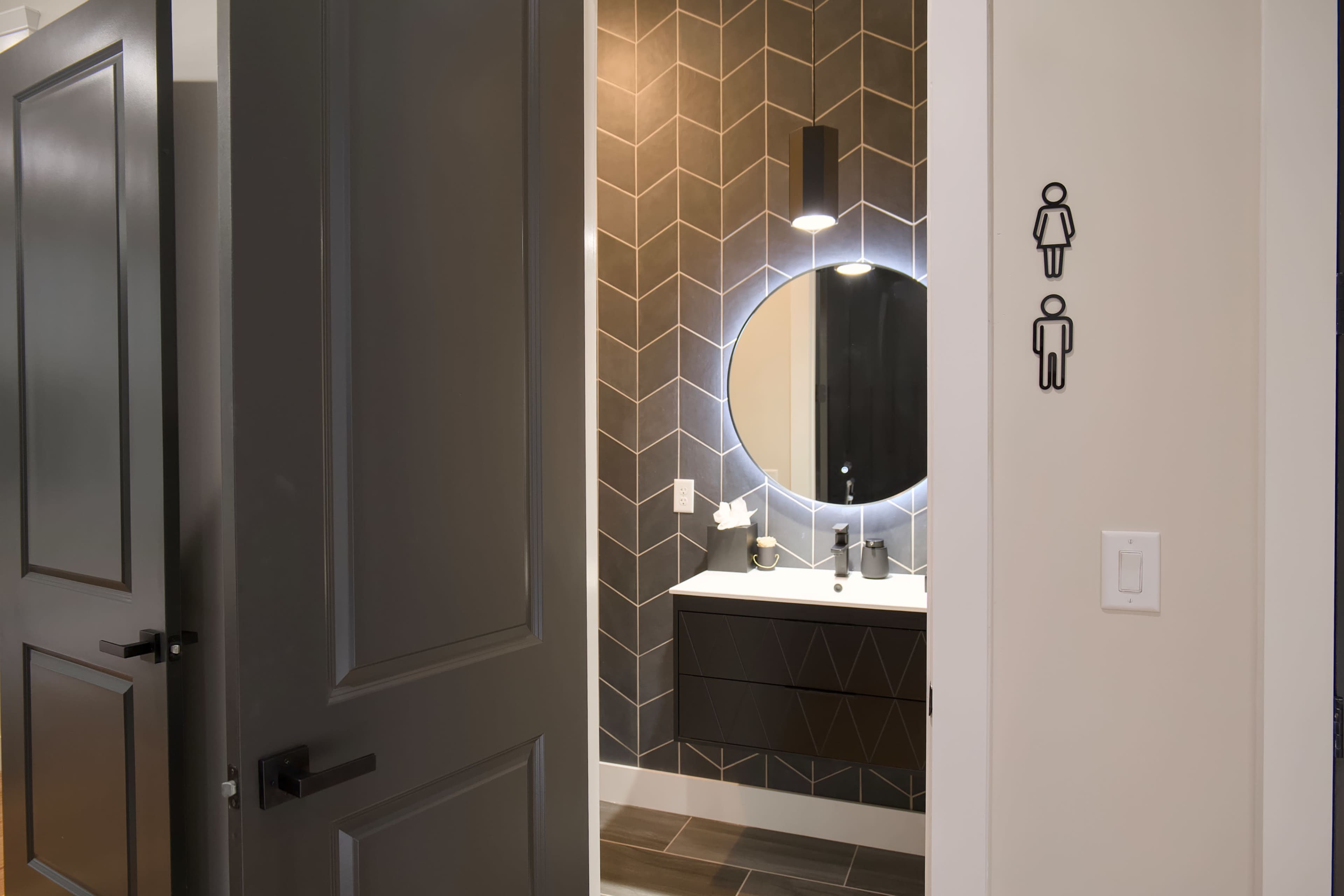 A well-lit bathroom features a round mirror and a dark geometric patterned wall, with gender symbols indicating restroom facilities nearby.