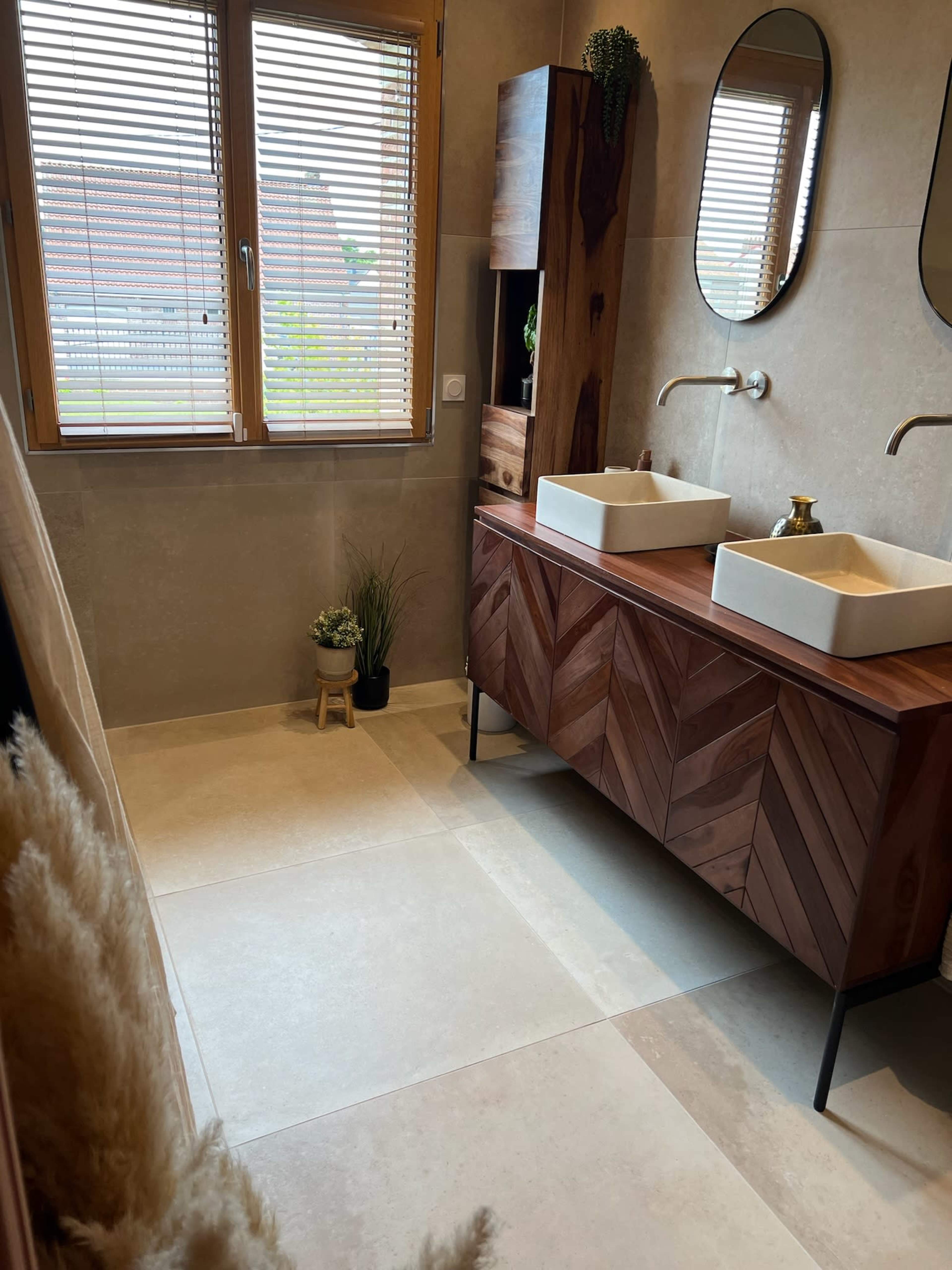 A modern bathroom featuring a wooden cabinet with two sinks, large mirrors, and a window with blinds.
