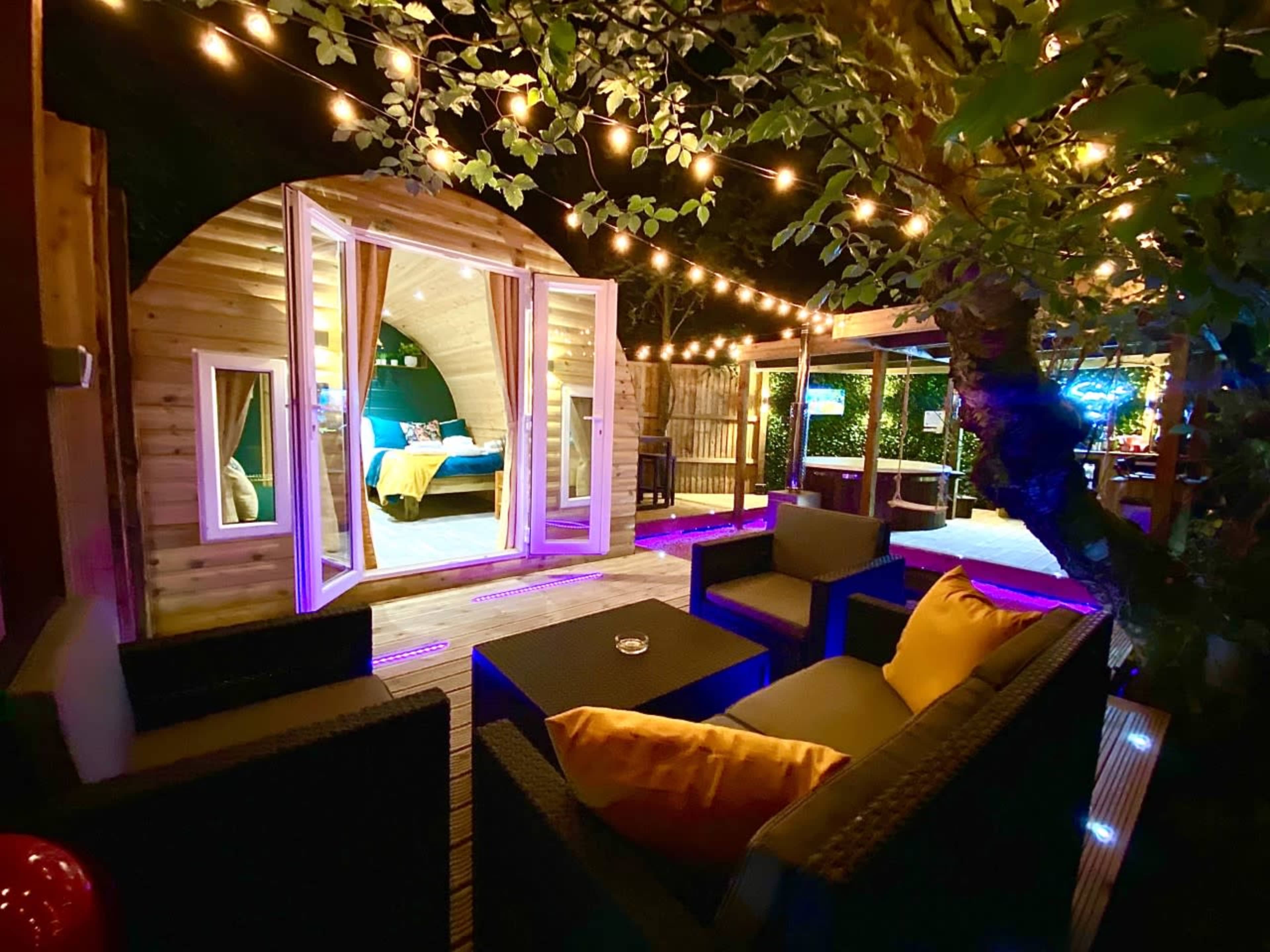 A cozy outdoor seating area is illuminated by string lights, leading to a unique wooden cabin with large windows and a neatly arranged interior.