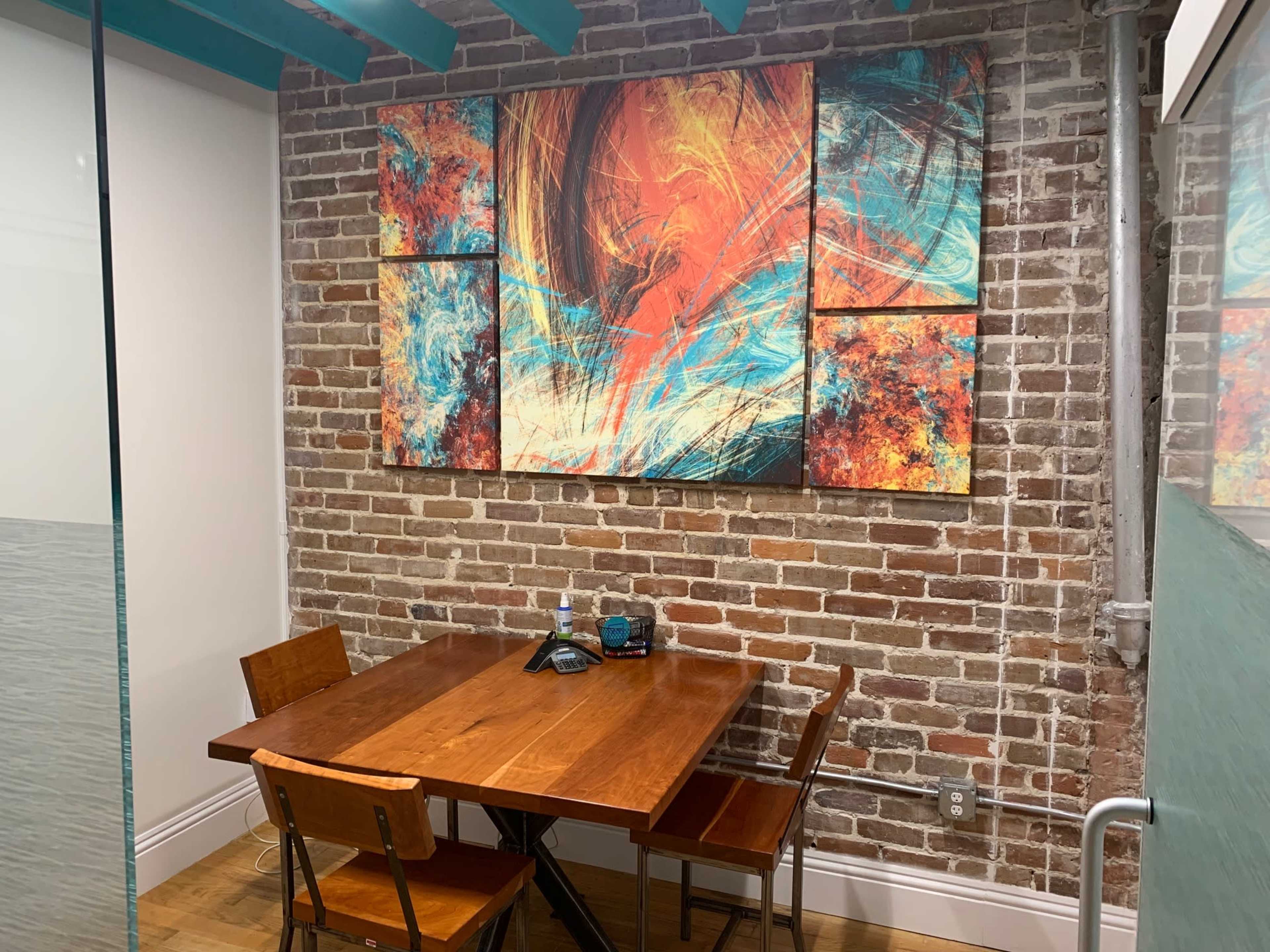 The image shows a small meeting room with a wooden table and three chairs, featuring a colorful abstract artwork on a brick wall.