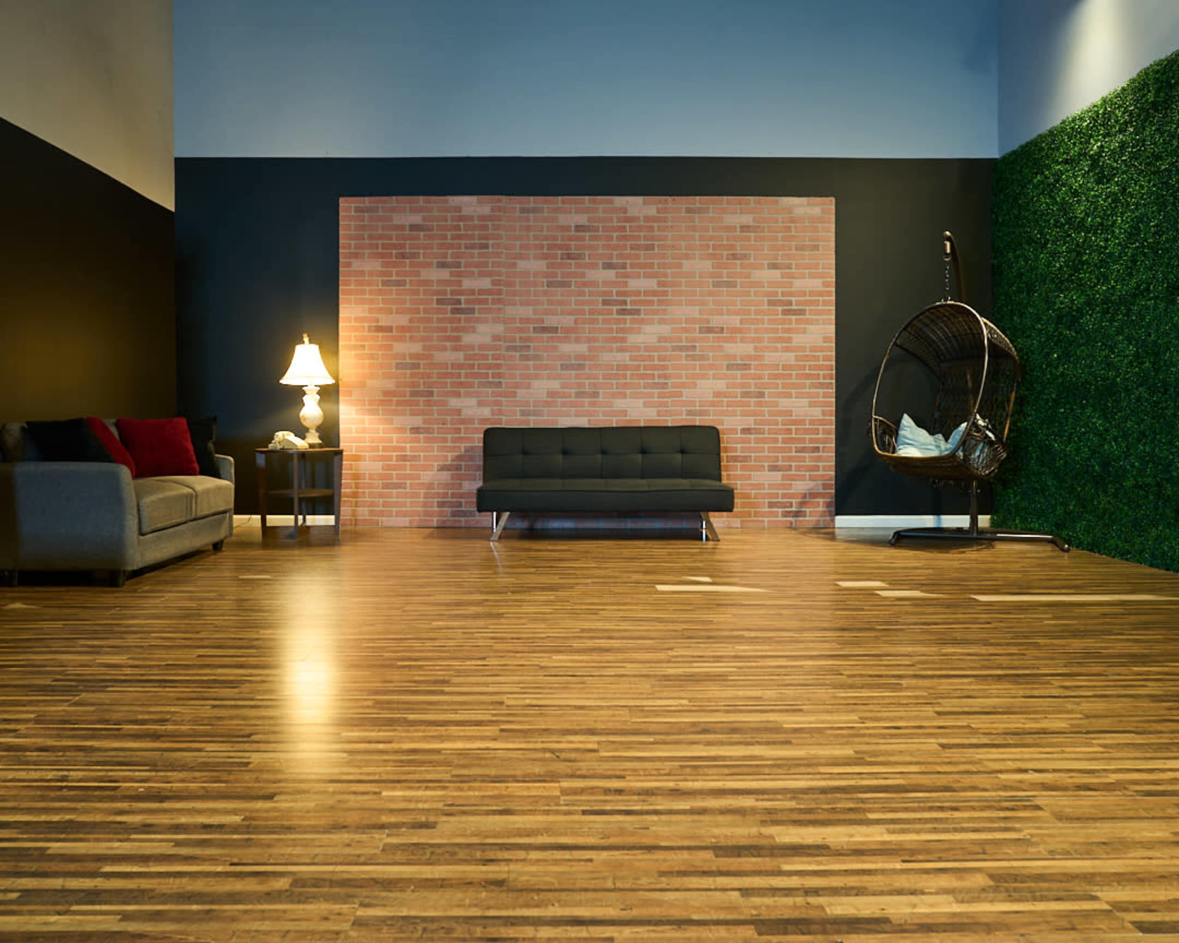 A spacious interior featuring a wooden floor, a gray sofa, a black couch, a hanging chair, a lamp, and a wall with a brick pattern and artificial greenery.