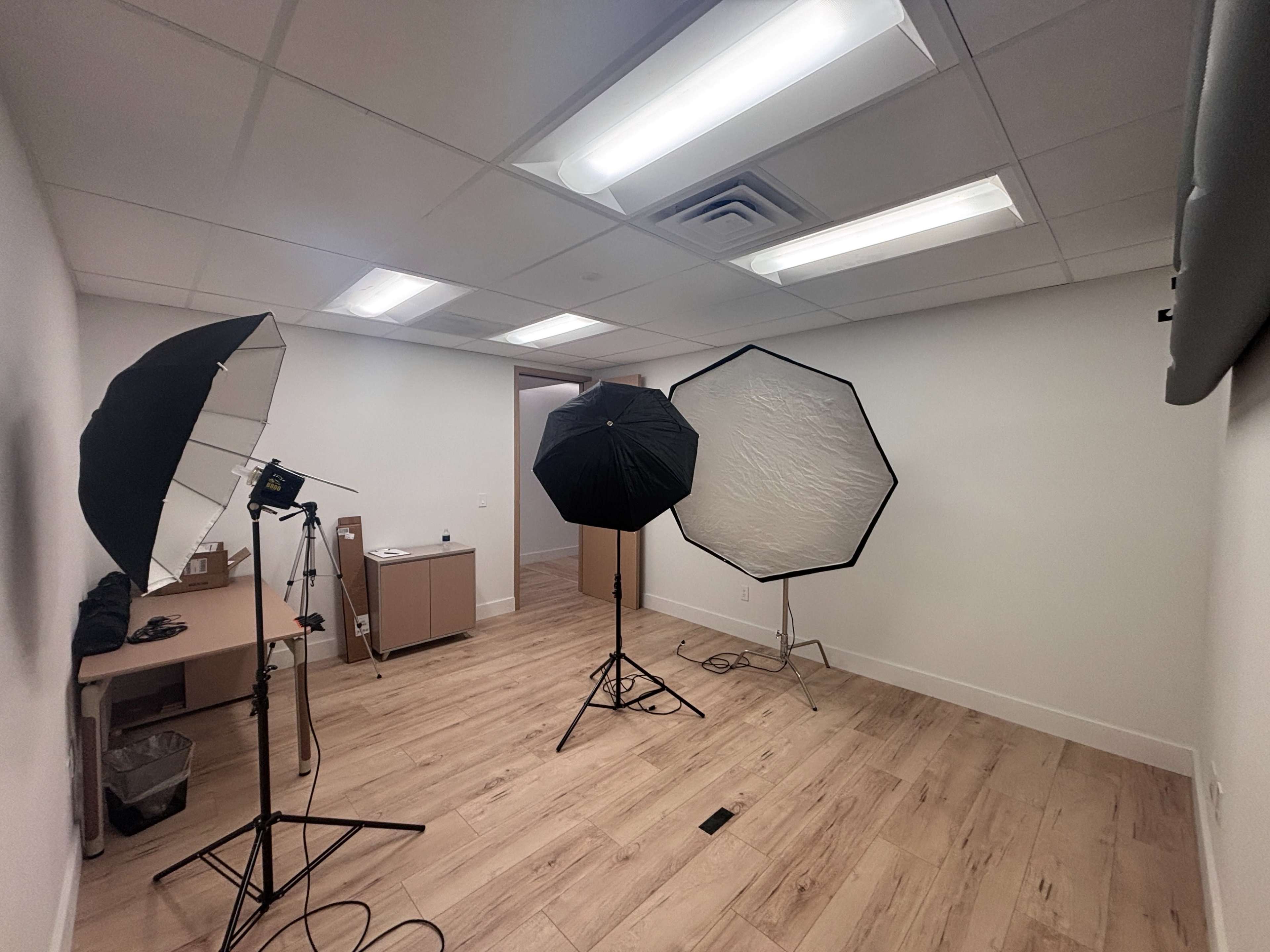 The image shows a photography studio equipped with two softbox lights and a black umbrella reflector, set up in a sparsely furnished room with light-colored walls and wooden flooring.