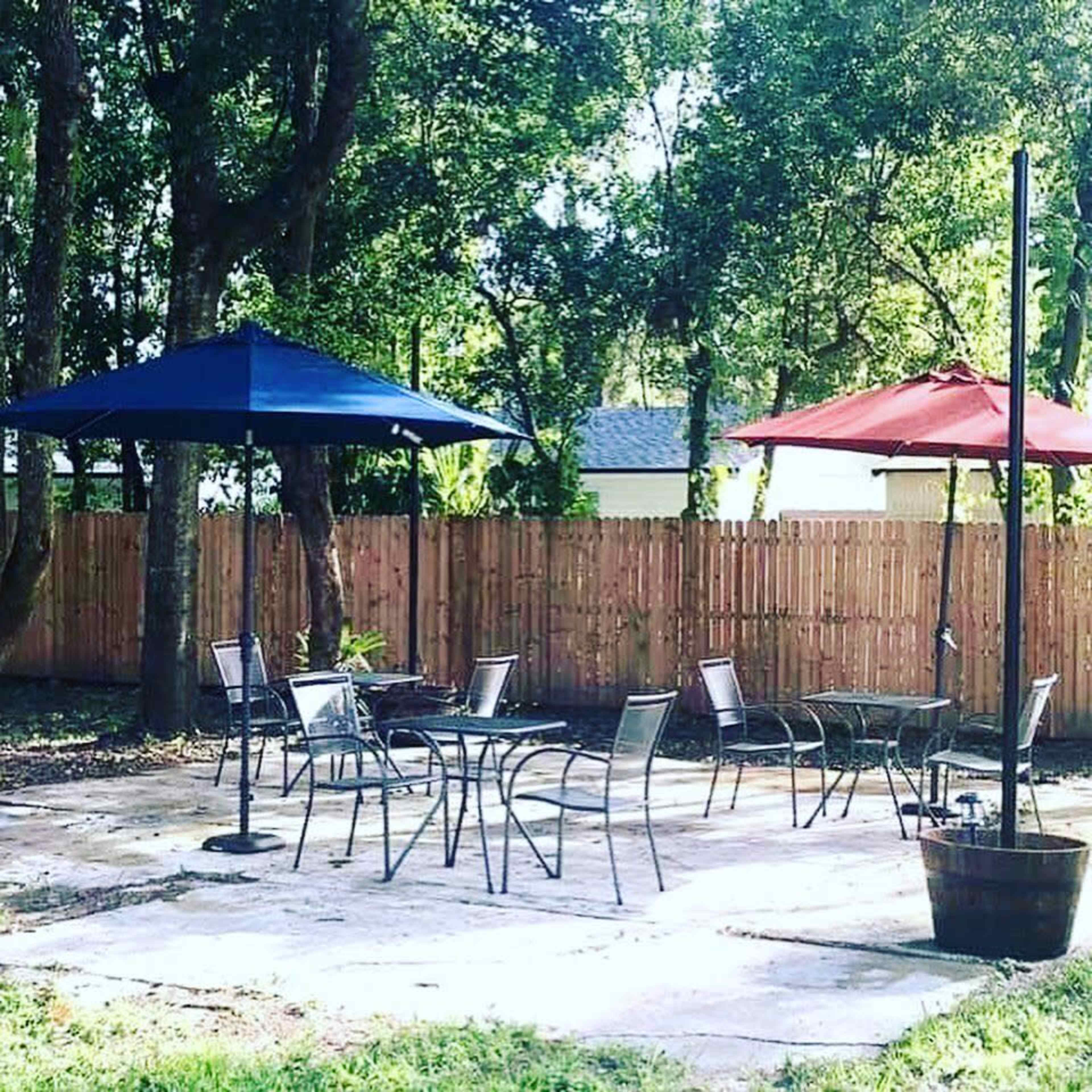 The image shows a patio area with several metal tables and chairs beneath blue and red umbrellas, surrounded by trees and a wooden fence.