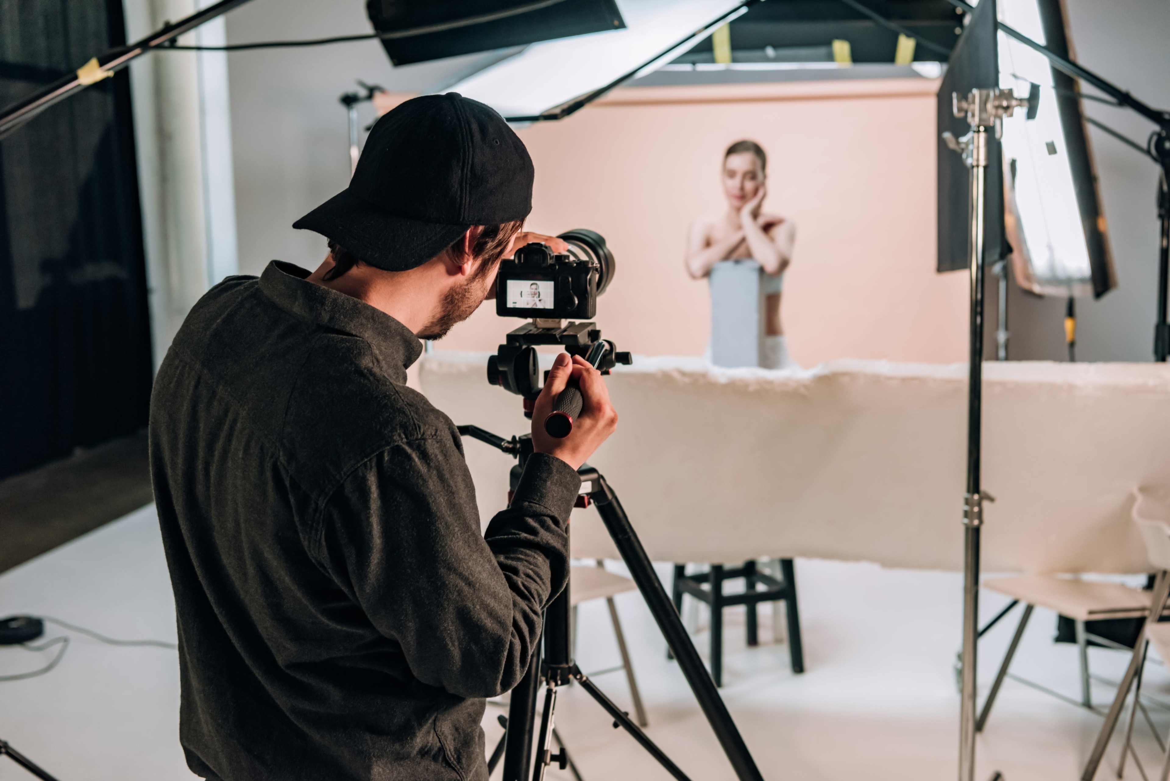 A photographer is using a camera on a tripod to capture an image of a model posing in front of a backdrop in a studio setting.