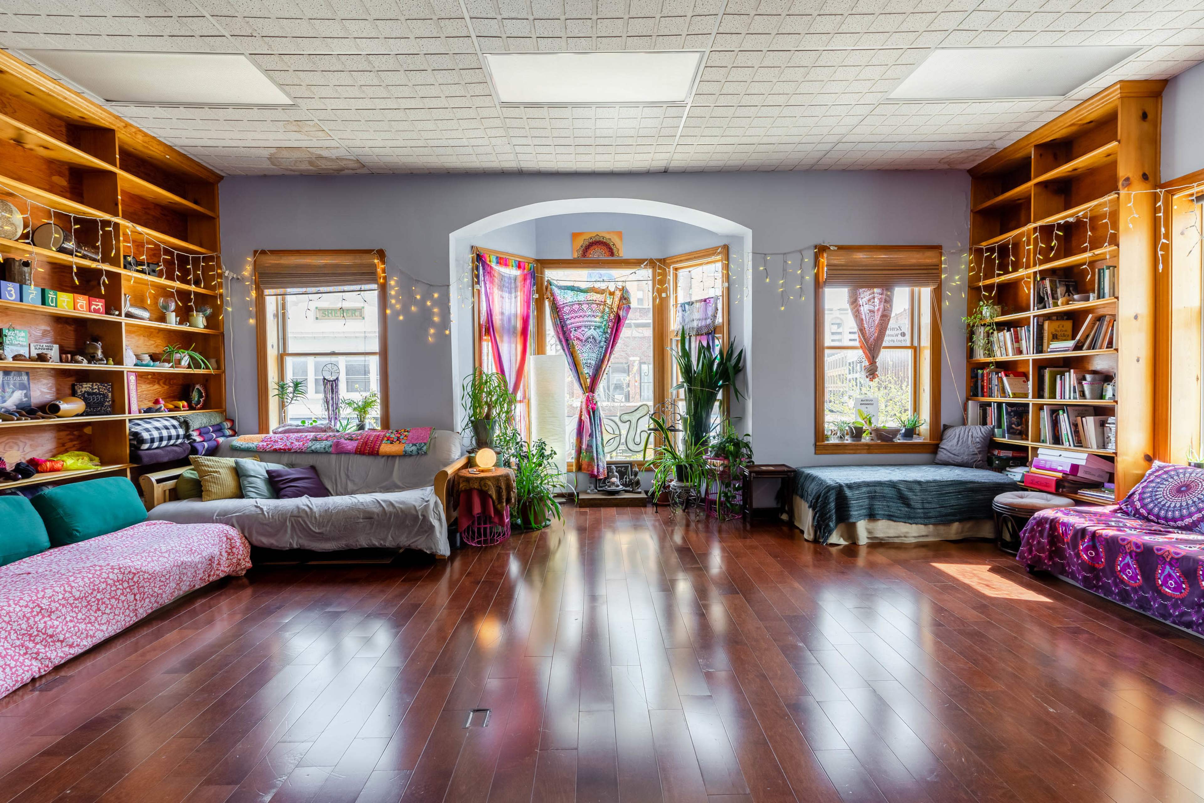 A spacious room with wooden shelves filled with books and colorful cushions arranged around large windows adorned with plants and fabric drapes.