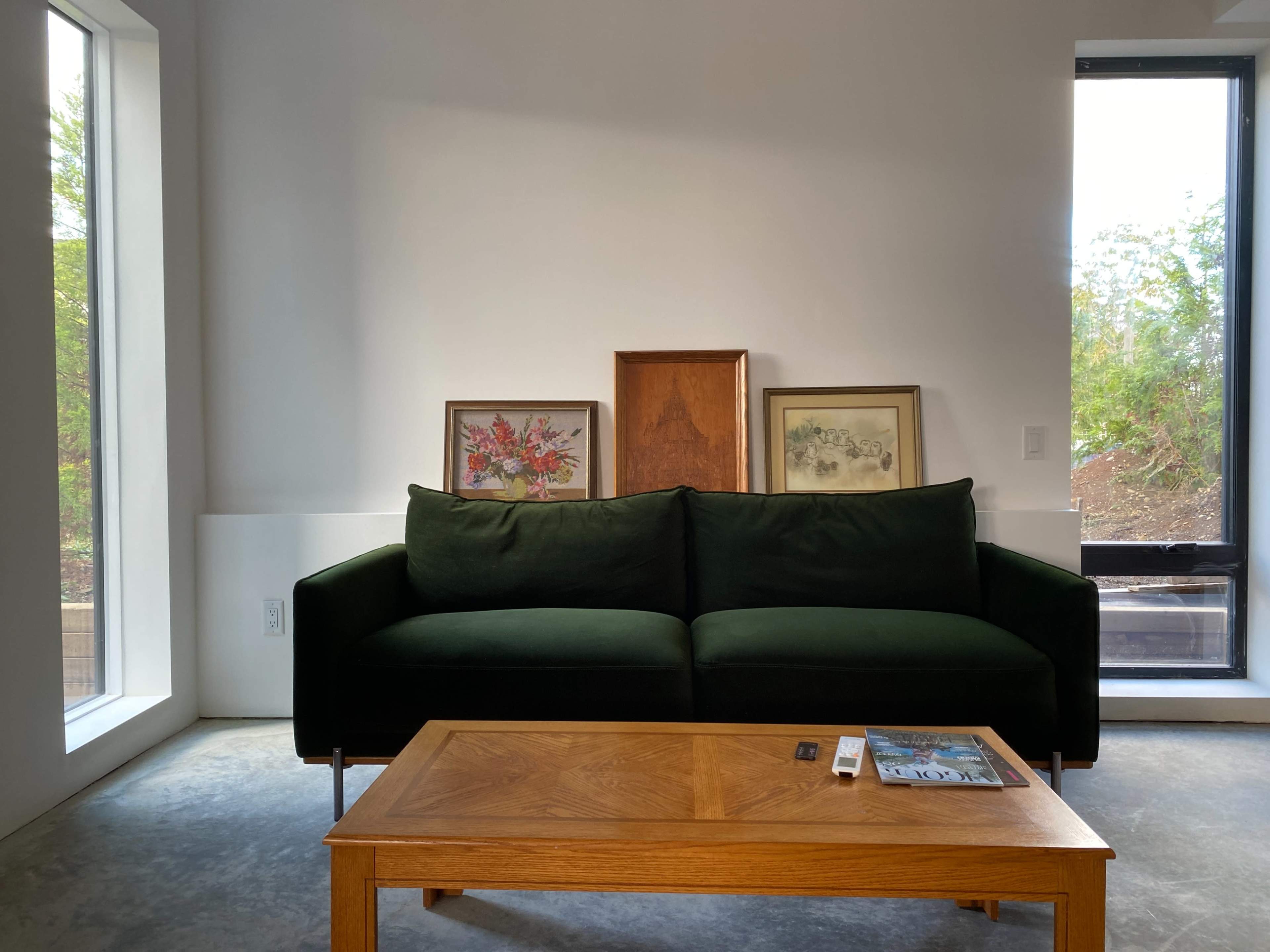 A green velvet sofa is positioned in a bright room, accompanied by a wooden coffee table and framed artwork on the wall.