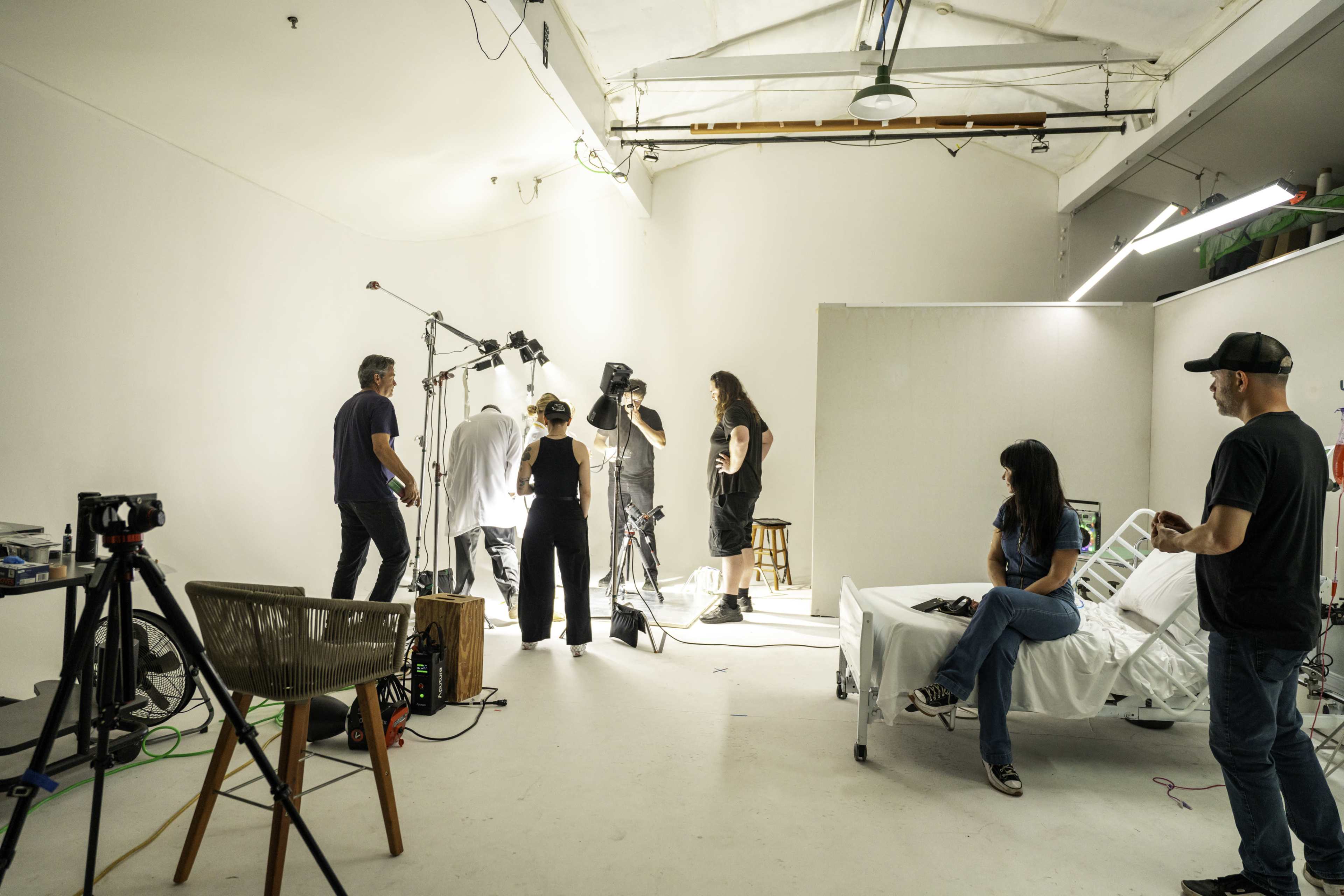 A film crew is setting up equipment in a brightly lit studio space featuring a hospital bed.