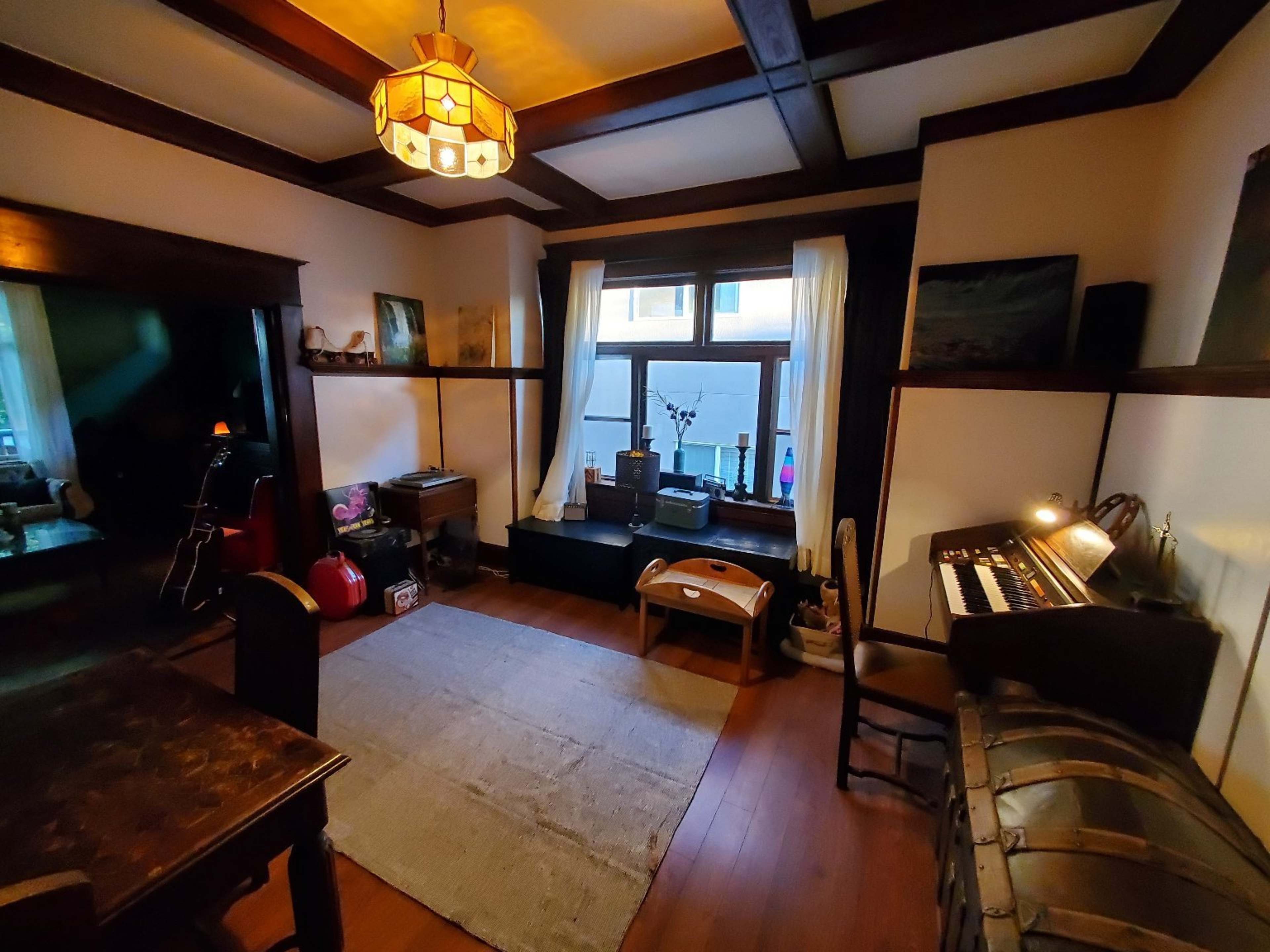 A cozy room with wooden beams, a vintage lamp, a window with sheer curtains, and various furnishings including a small table and an organ.