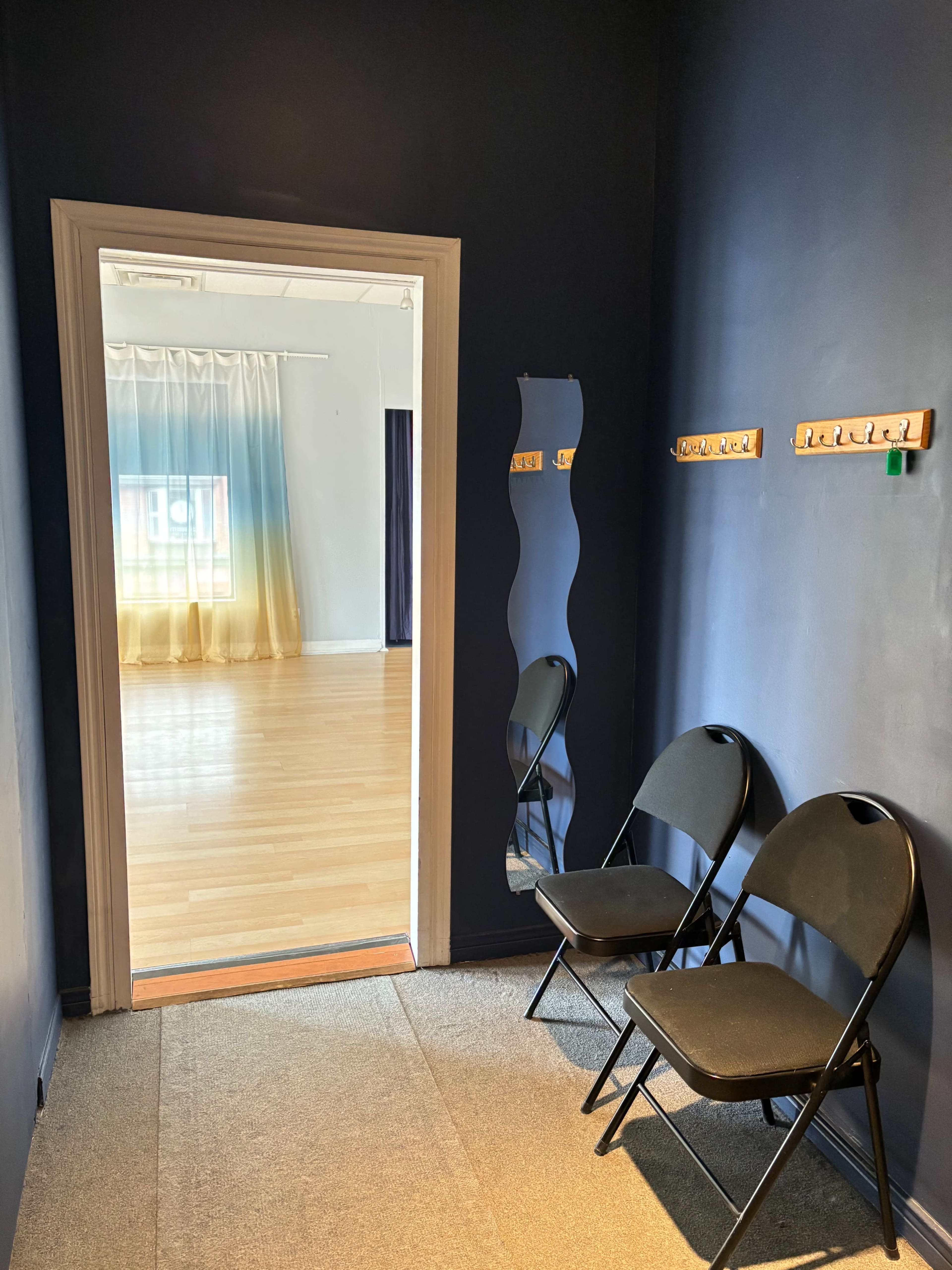 The image shows a small waiting area with two black folding chairs near a doorway leading to a bright room with large curtains and wooden flooring.