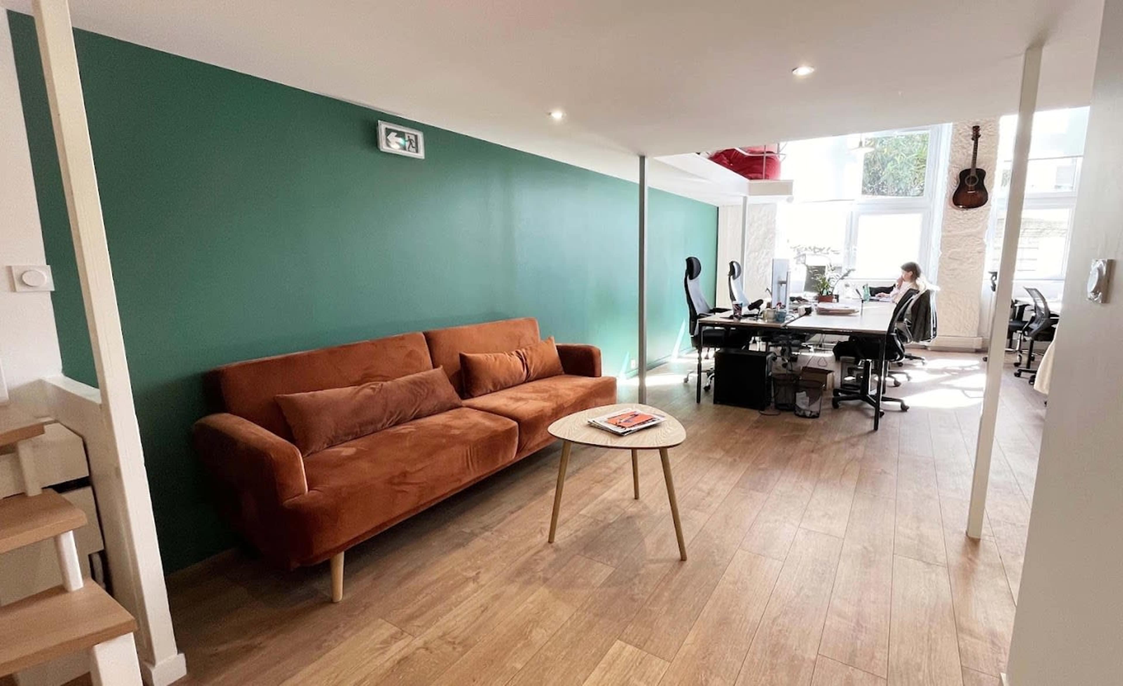 The image shows a modern living space featuring a brown sofa, a small round table, and a workspace with desks and a chair against a green wall.