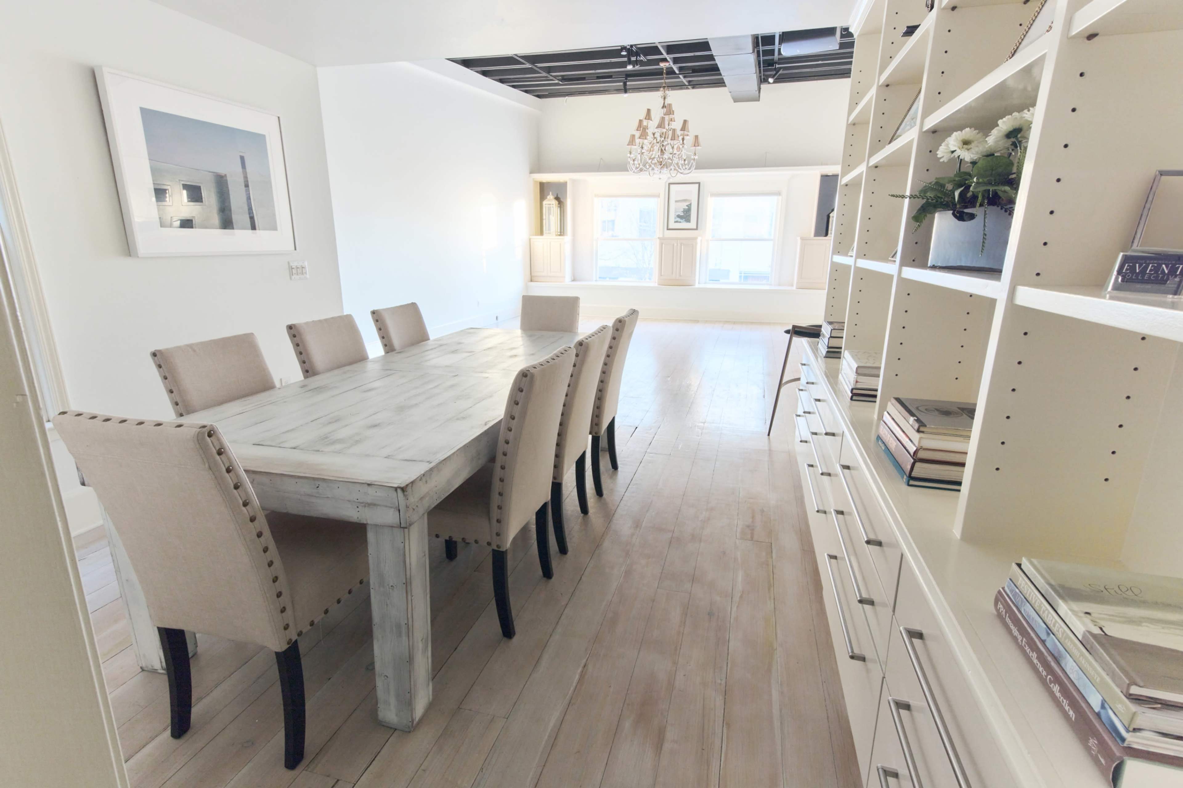 The image shows a dining area with a long wooden table surrounded by beige chairs, leading to a bright, open living space with built-in shelves and a chandelier.
