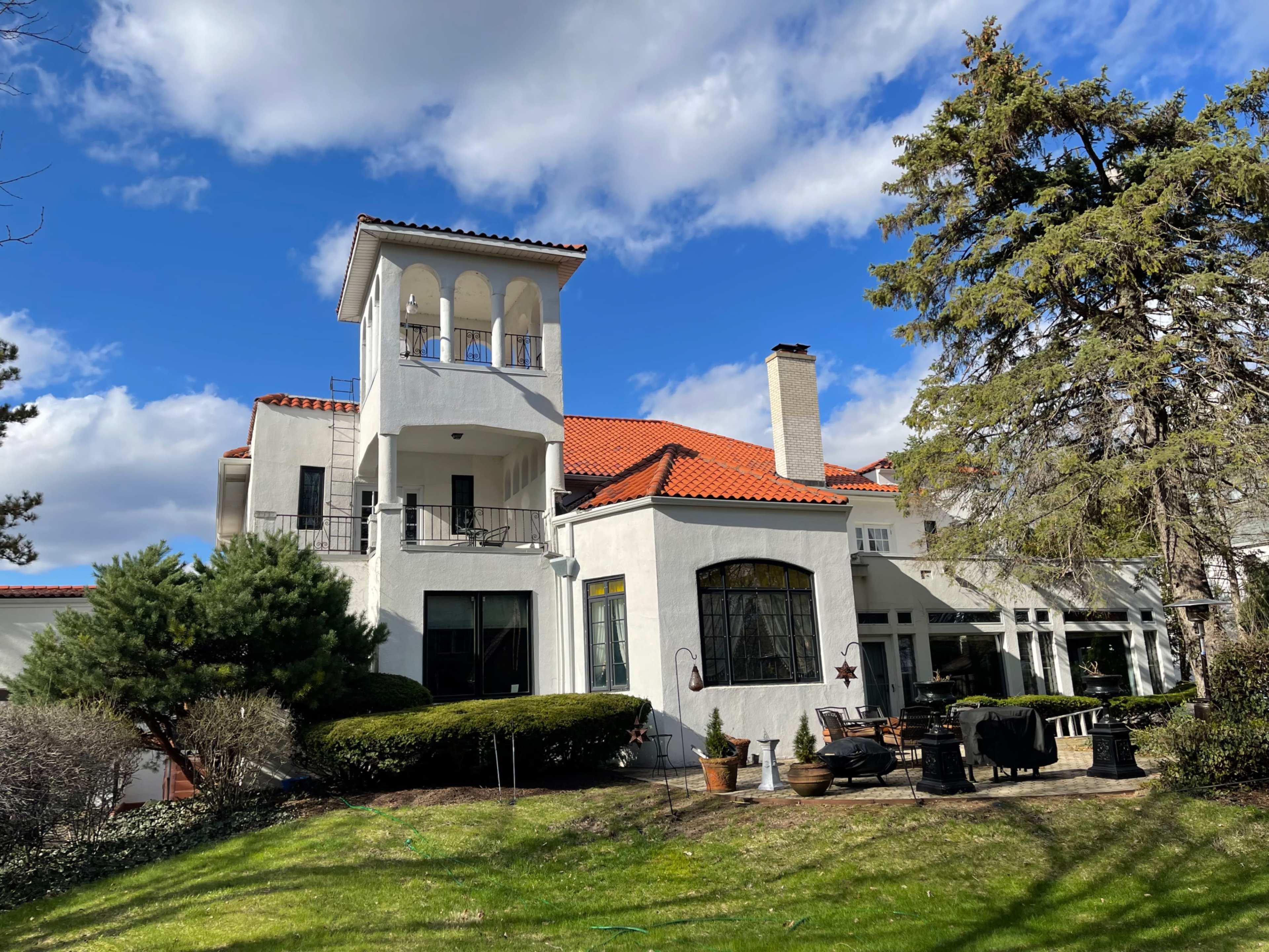 A large, two-story white house with a red tile roof and a tower overlooks a manicured lawn and garden.