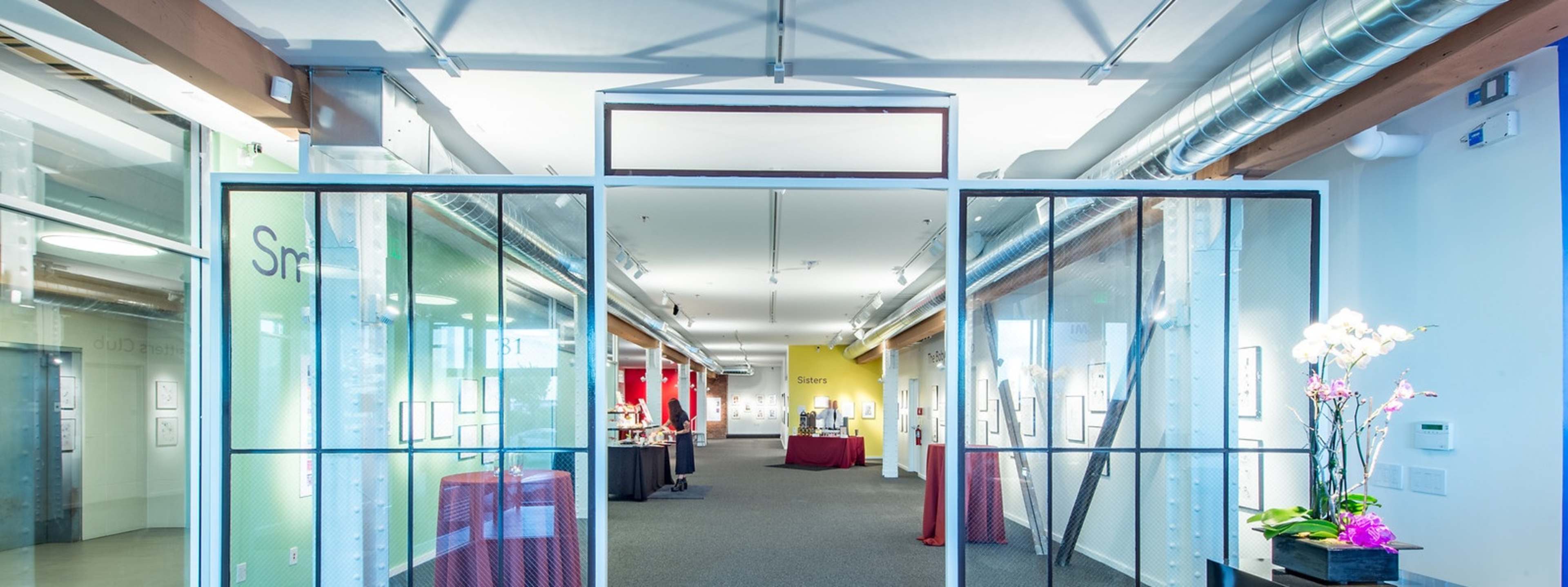 A modern art gallery with large glass doors leading into a well-lit space featuring artwork displayed on the walls and tables set up for an event.