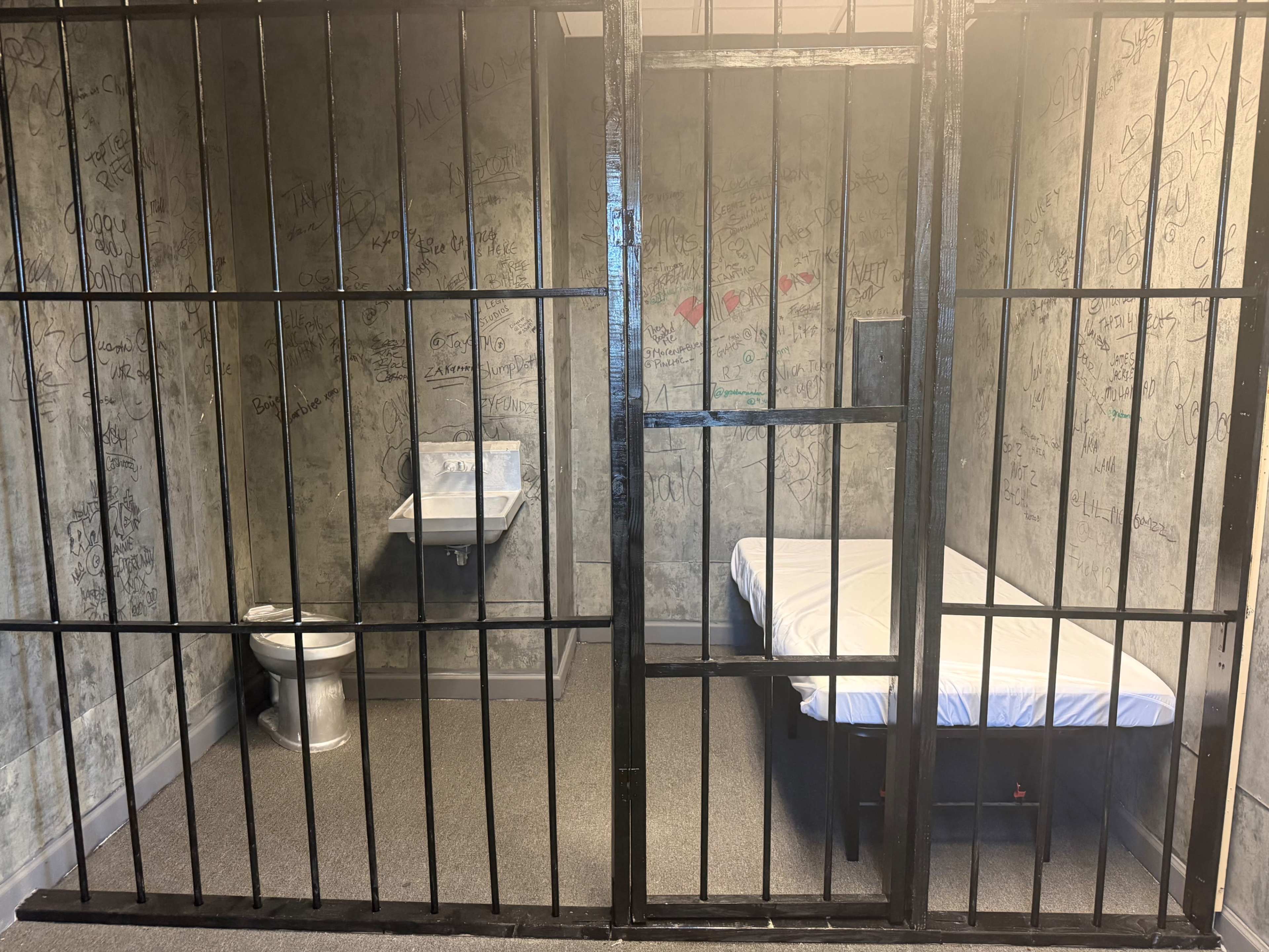 The image shows the interior of a prison cell with a bed, a toilet, and walls covered in graffiti.