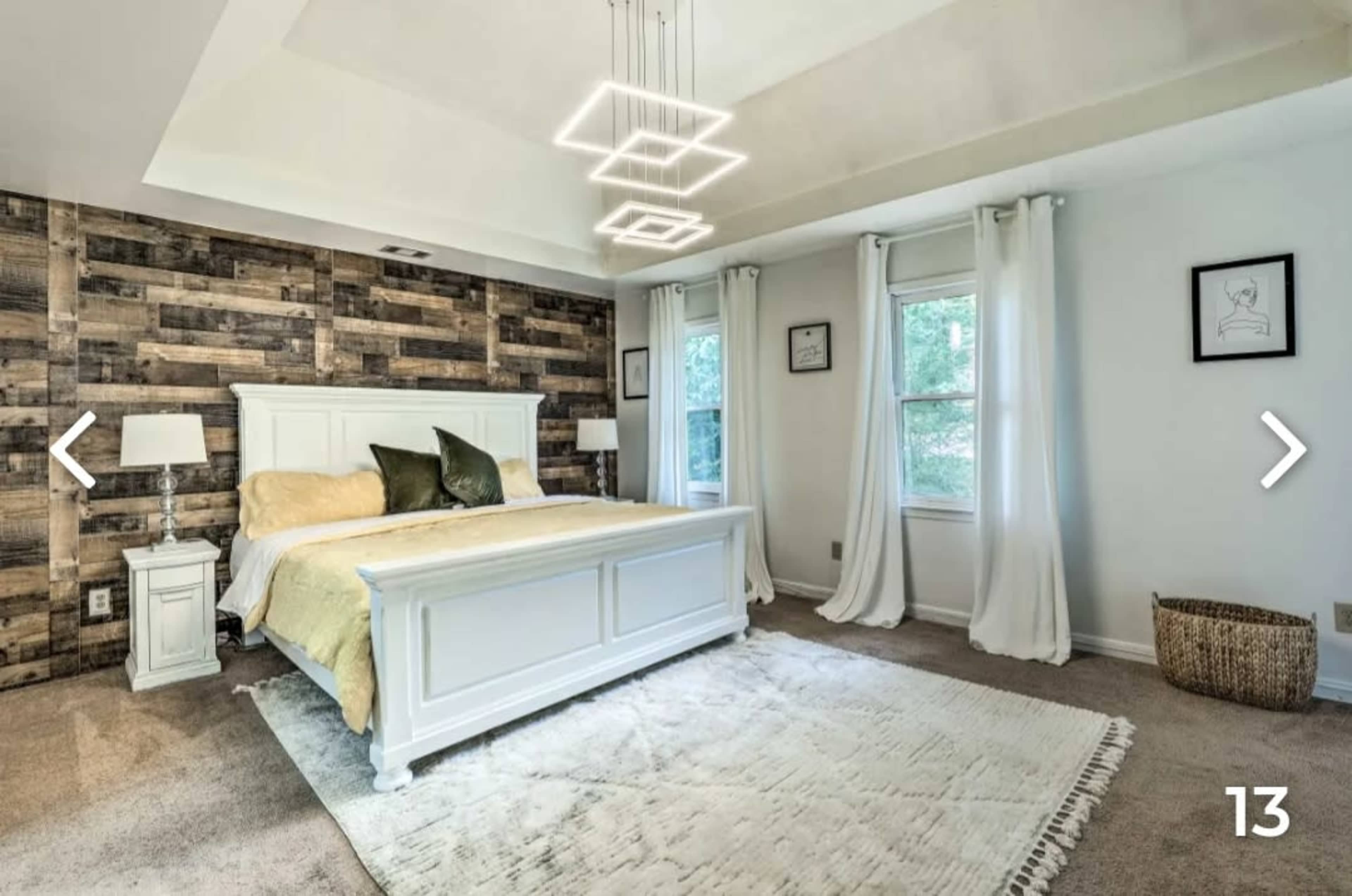 The image shows a bedroom featuring a white bed with a wooden accent wall, large windows with curtains, and a rug on carpeted flooring.