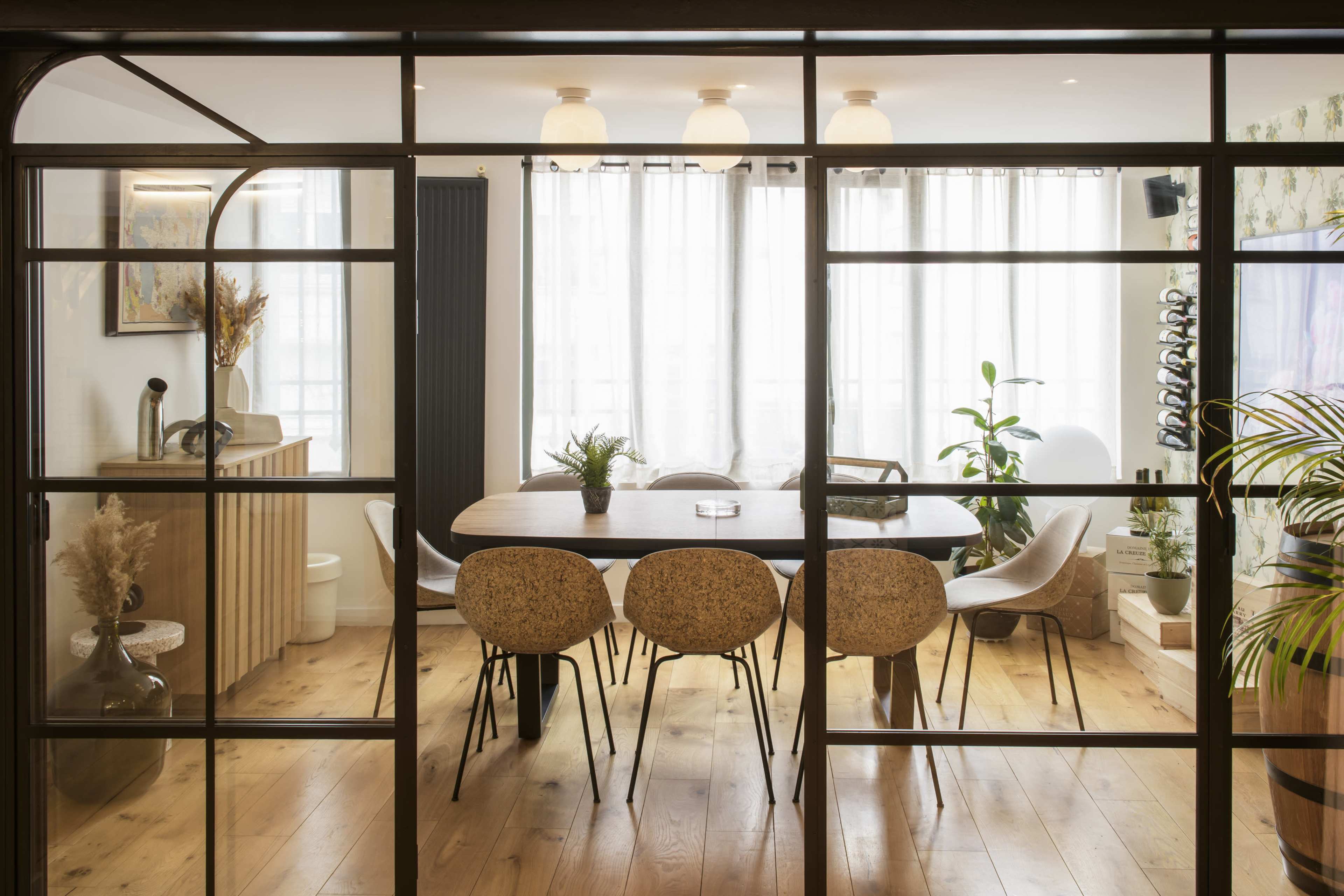 A dining area features a large wooden table surrounded by various styled chairs, illuminated by natural light streaming through sheer curtains.
