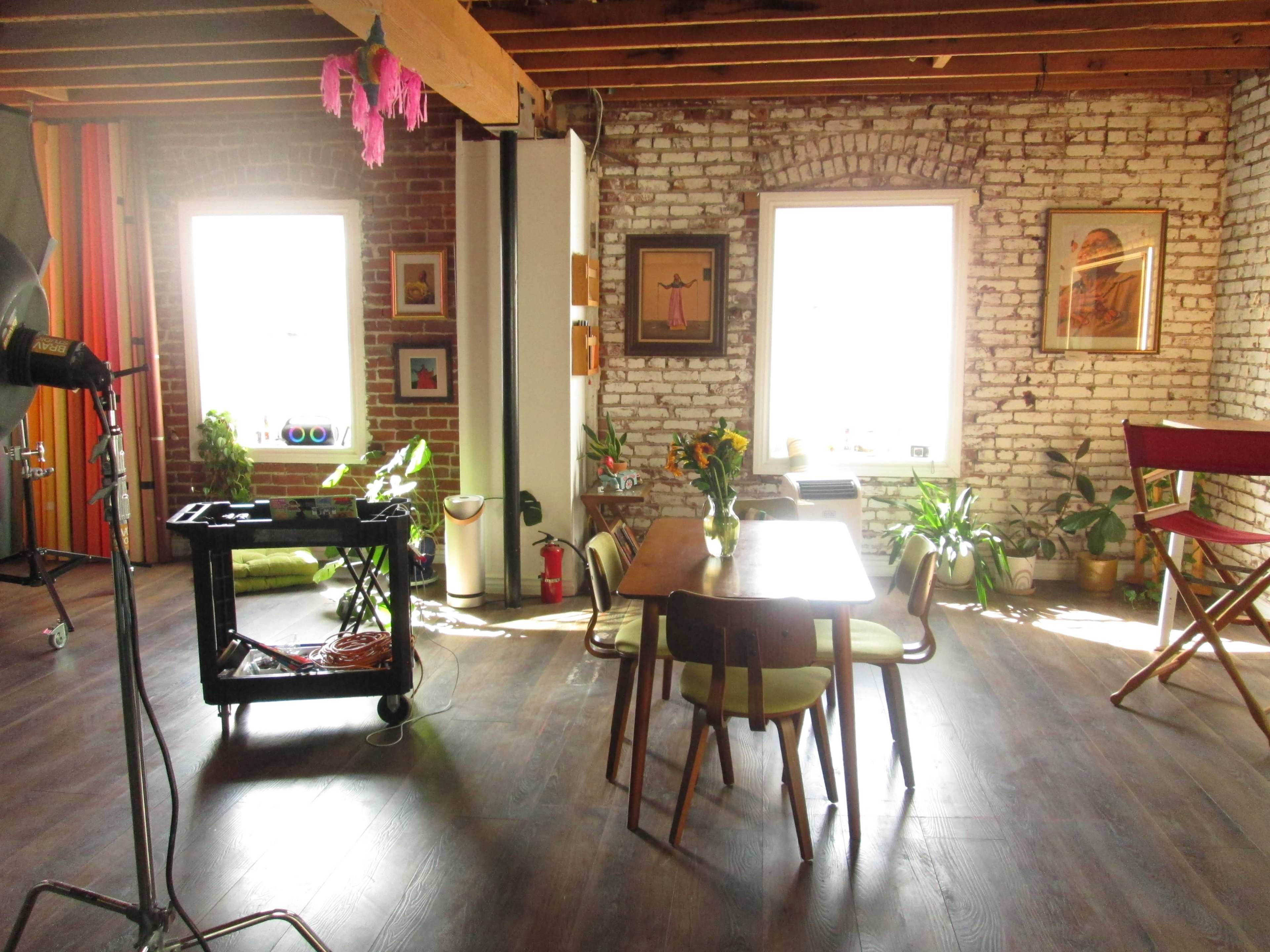 The image shows a brightly lit room with exposed brick walls, featuring a wooden dining table surrounded by chairs and several potted plants.