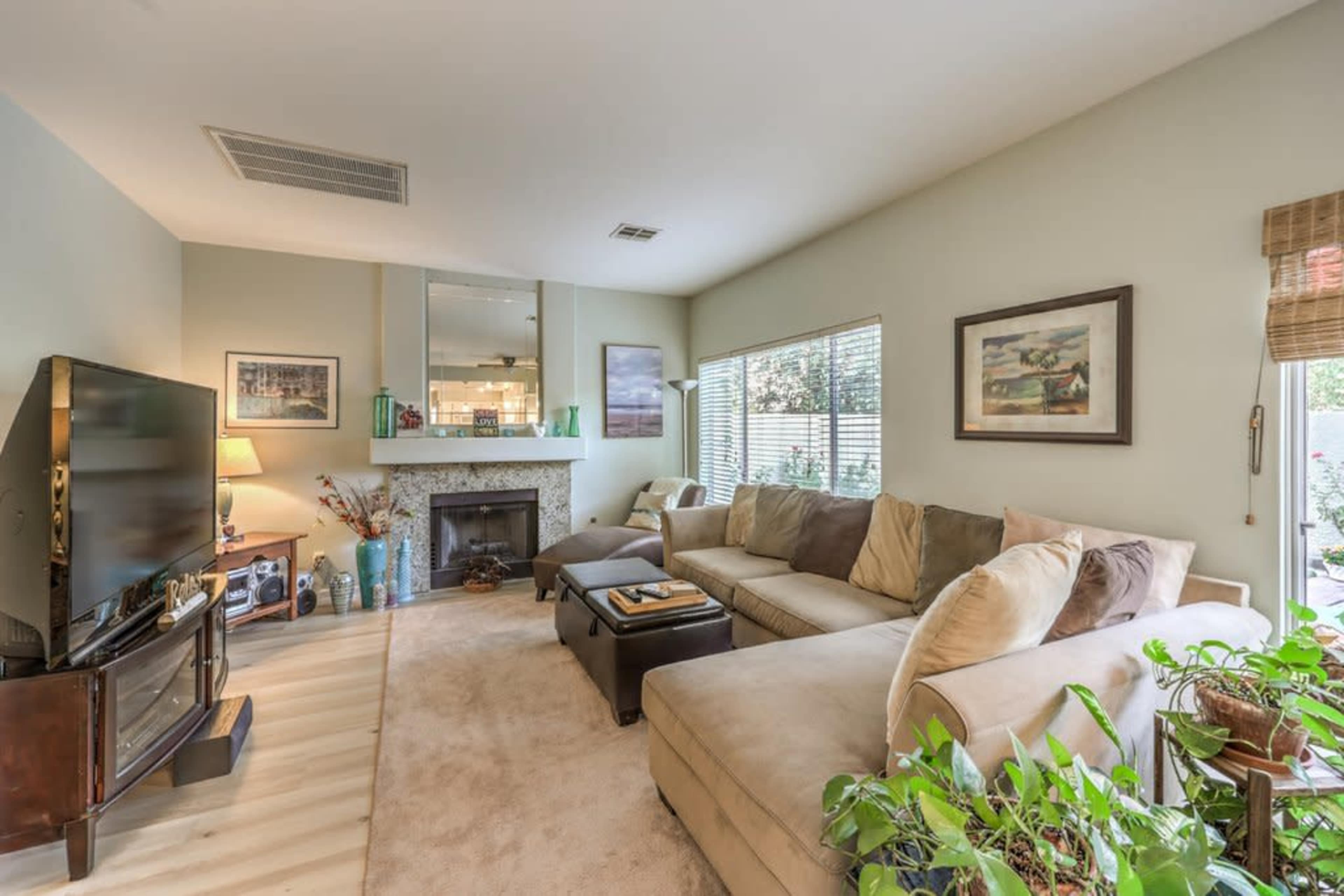 A living room features a sectional sofa, a television, a fireplace, and several decorative plants in the corners.